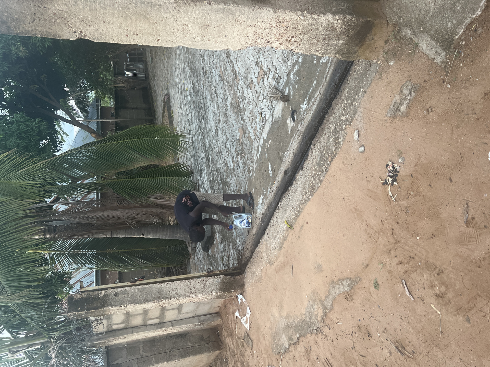 A man in a black t-shirt and patterned shorts is pouring oil from a white container onto a paved area next to a curb. The location appears to be outdoors, with a palm tree and other foliage nearby. There is evidence of water on the paved surface, and the adjacent ground is sandy and uneven. The location might be in Makurdi, Nigeria, but that cannot be forensically confirmed from the image.