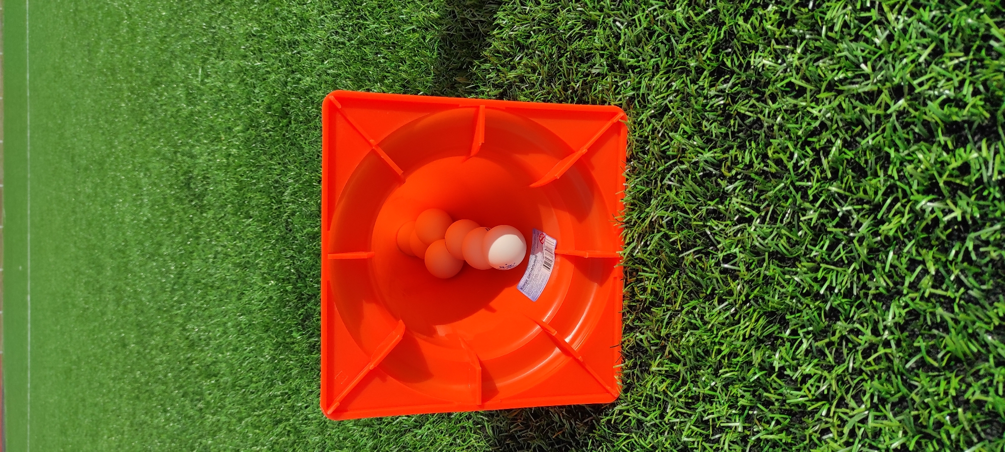 An orange traffic cone is placed on artificial green turf. Inside the cone, a string of white ping pong balls is visible, with seven balls arranged in a line. The cone has a label on its inner surface, but the text is not clearly legible. The background shows more of the green turf and a white line marking the edge of what appears to be a sports field. The lighting suggests it is daytime, with shadows cast by the cone. The image appears to be taken from a slightly elevated perspective, looking down into the cone.