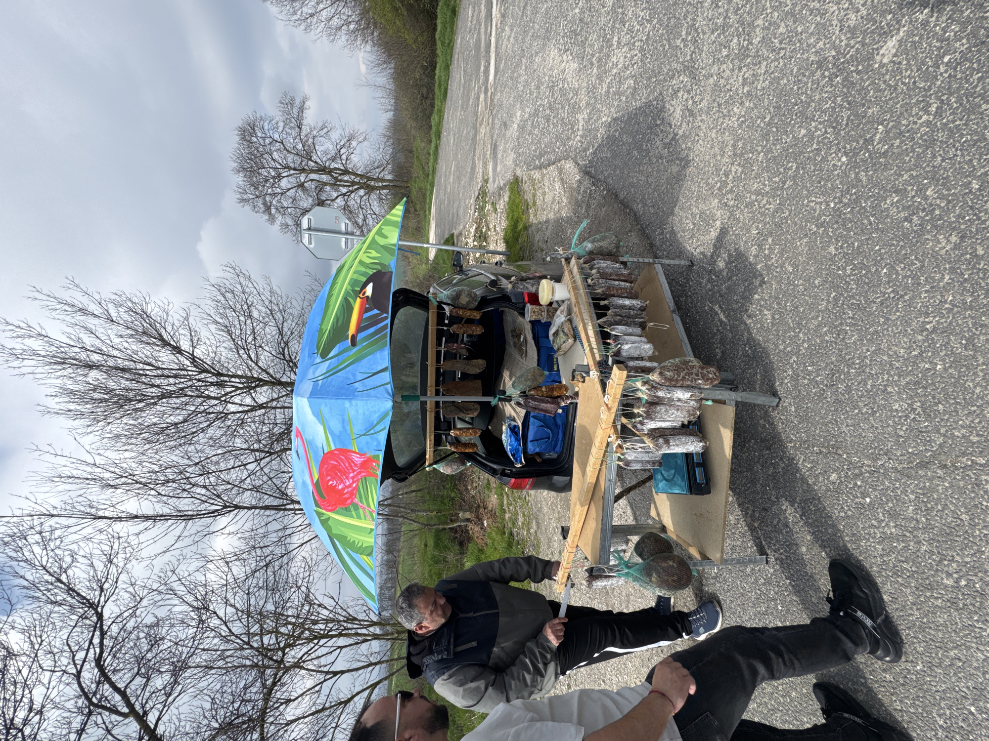 The image shows a roadside stall selling sausages in Bliznatsi, Bulgaria. The stall is set up next to a car with its trunk open, using a makeshift frame to hang the sausages. A blue umbrella with images of a toucan and flamingo provides shade. A man is seen handling the sausages, while another man is partially visible in the foreground. The scene is set on an asphalt road with vegetation and trees in the background under a cloudy sky.