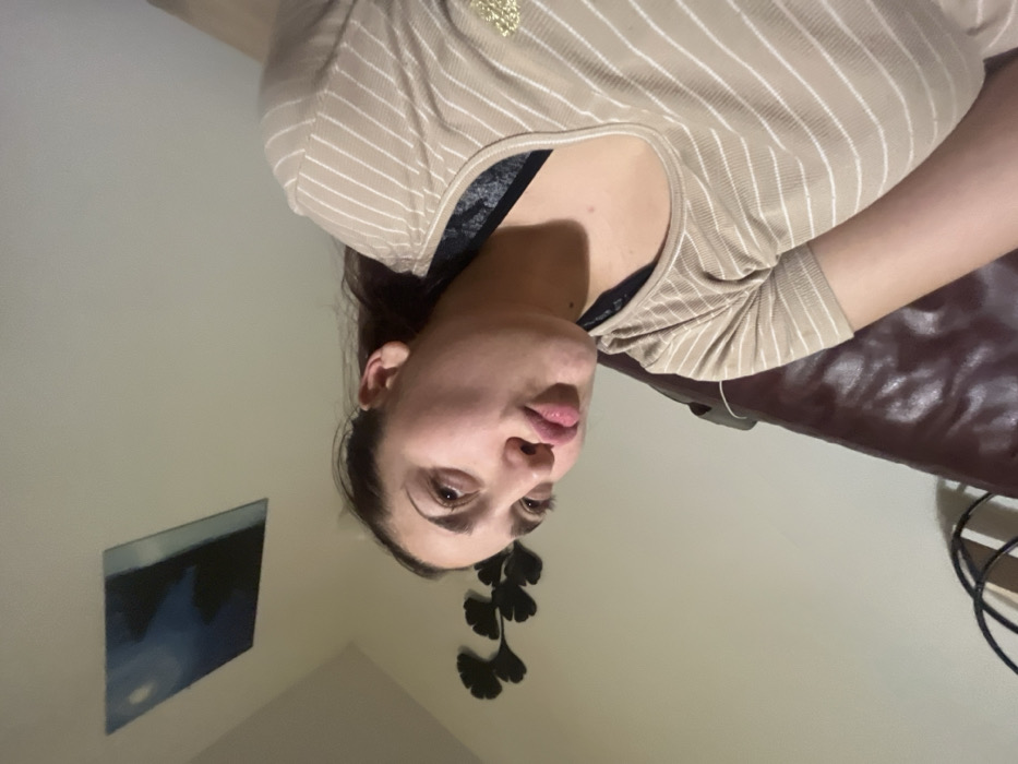 A woman is seen from a low angle, wearing a beige striped top with a black underlayer peeking out. Her face is slightly tilted towards the camera, revealing her eyes and lips. Behind her, a dark leather couch is partially visible. The wall features a framed painting with blue hues and a dark, stylized leaf-shaped decoration. The scene has indoor lighting, and the image is rotated, giving a slightly disorienting perspective.
