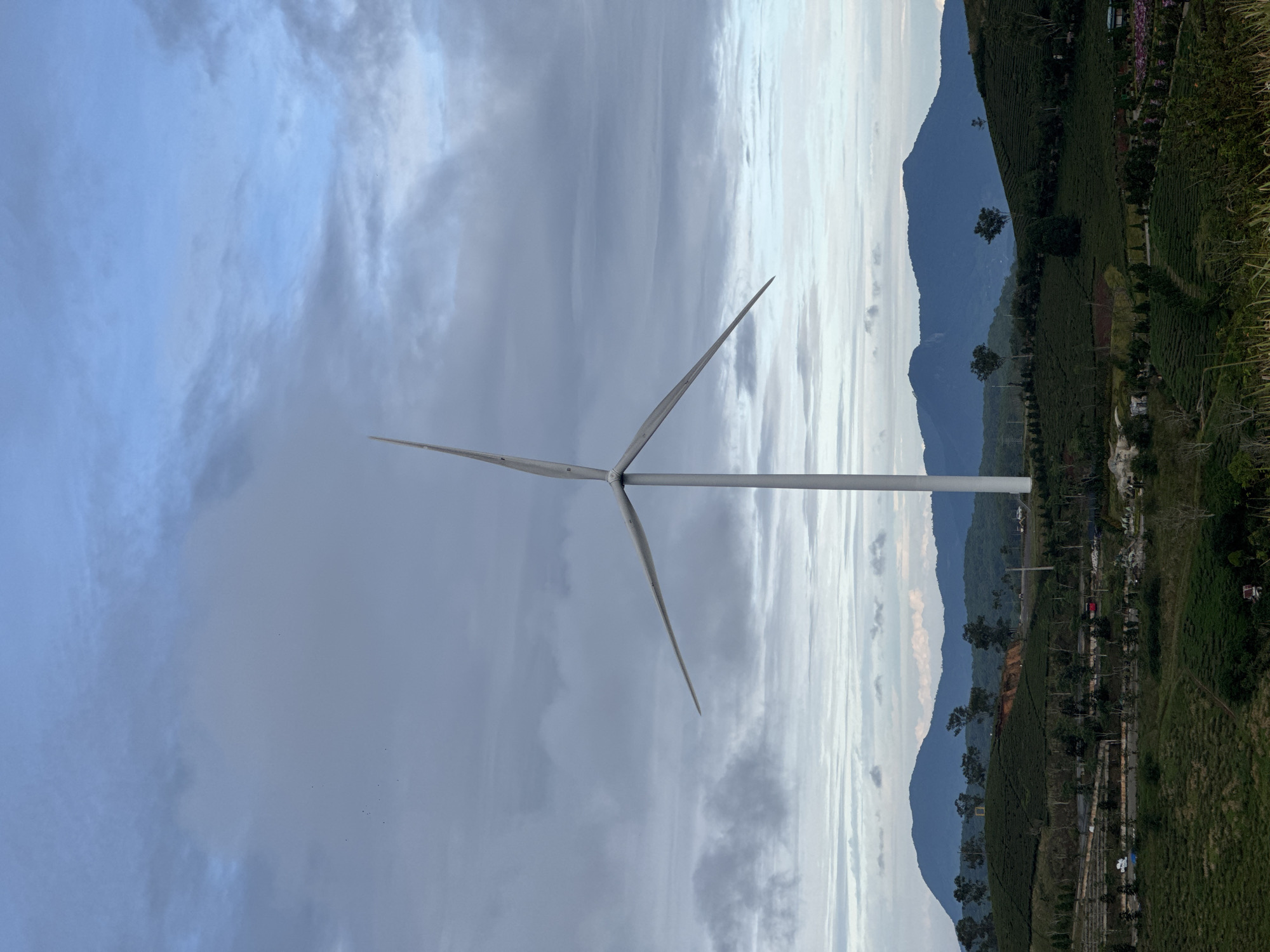 The image shows a wind turbine in Phát Chi, Vietnam. The turbine is white with three blades and stands tall against a backdrop of cloudy skies. In the distance, mountain ranges are visible, and the land around the turbine appears to be lush with green vegetation and tea plantations. The photograph is taken from a slightly low angle, emphasizing the height of the turbine.