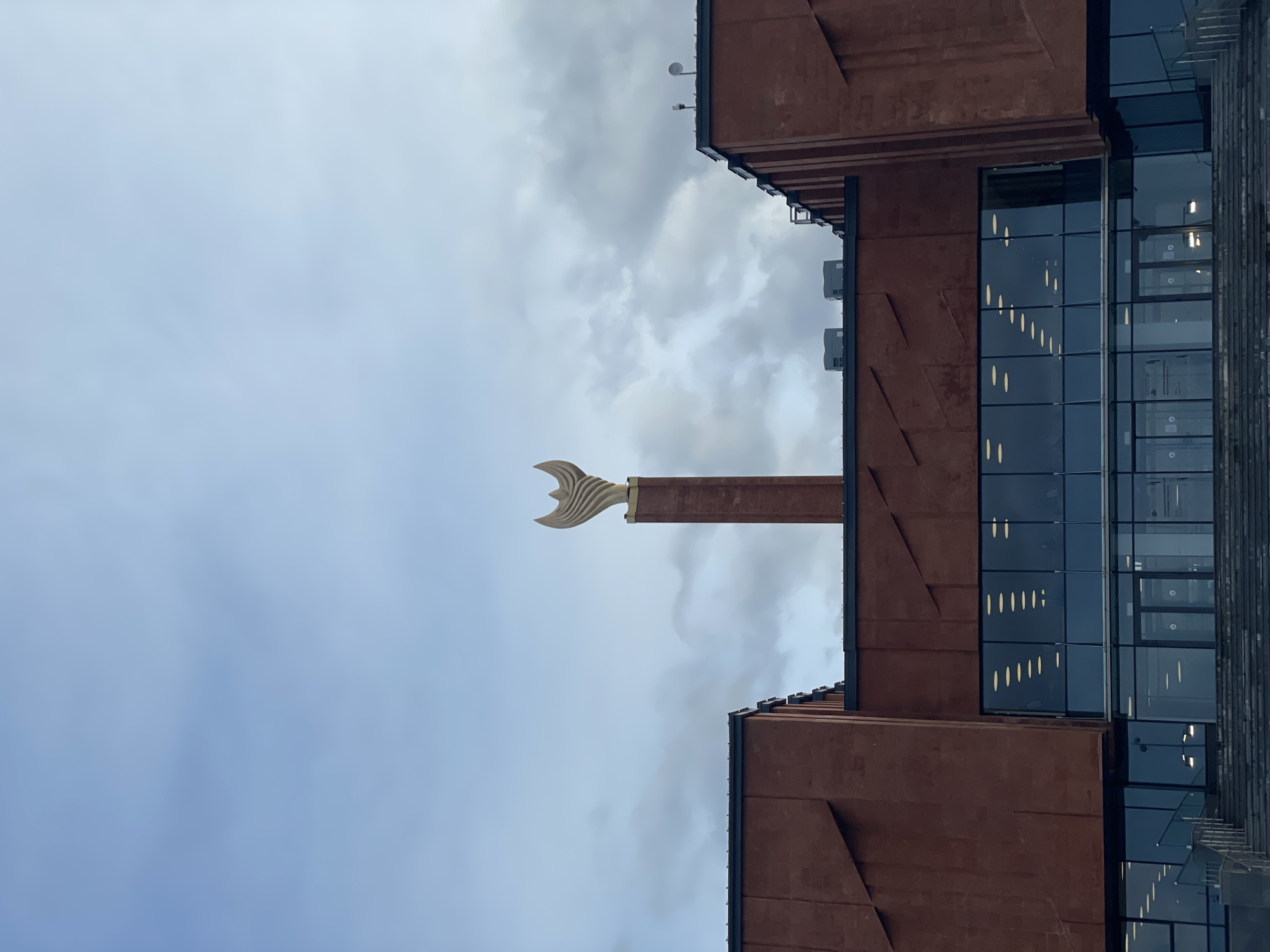 A modern architectural scene captured on an overcast day features a striking building and a unique sculpture. The image, oriented vertically, showcases a large building with a reddish-brown facade, possibly constructed from corten steel or a similar material. This building exhibits distinct vertical and horizontal structural elements, with several large sections of dark glass windows reflecting scattered interior lights, suggesting a contemporary design.
Extending upwards from the building's structure is a tall, narrow column, topped by a prominent, golden, abstract sculpture. The sculpture is stylized, resembling a graceful, uplifted bird's wing or a fin, shining brightly against the muted sky.
The sky is overcast with soft, light grey clouds, providing a diffused natural light that highlights the textures of the building and the gleam of the sculpture. No people are visible, and no specific activity is taking place; the image is a static portrayal of the architectural subjects. The distinctive sculpture strongly resembles a modern interpretation of the Liver Bird, a symbol associated with Liverpool, United Kingdom, suggesting the location could be the University of Liverpool's School of Management building. No visible text is present in the image.