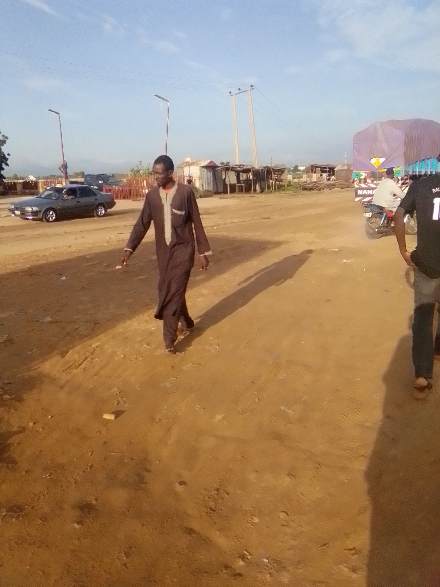 An outdoor scene in Gombe, Nigeria, shows a dirt road with a man walking towards the right foreground. He is wearing a brown traditional garment. In the background, there are several vehicles, including a dark sedan on the left, a motorcycle with a rider, and a large truck covered with a purple tarp. Structures resembling market stalls or temporary buildings are also visible. The ground is dusty and uneven, with tire tracks visible in places. The sky is clear and blue, suggesting it is daytime.