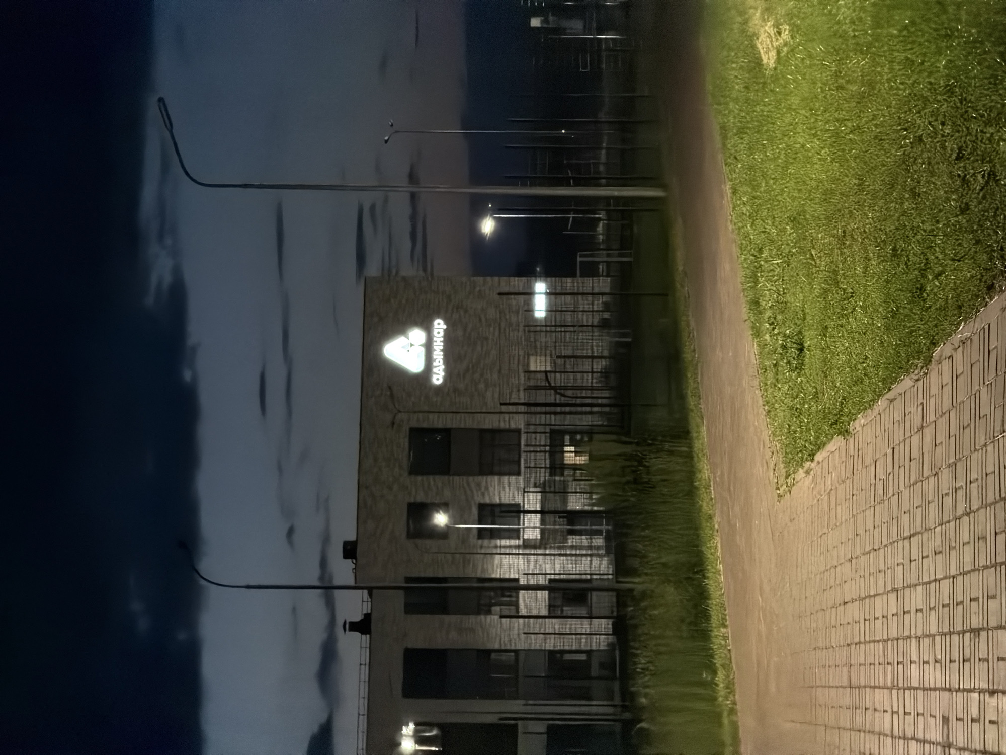 An exterior, nighttime, eye-level, medium shot shows a modern, brick building with a large, illuminated logo in white on its facade. The logo is a stylized triangle with a smaller triangle inside it, and the Cyrillic text "адымнар" is positioned below it. The building appears to be a school or community center, with multiple windows visible, some illuminated from within. The foreground features a paved walkway made of rectangular bricks, bordered by a strip of green grass and then taller green vegetation. In the background, tall metal structures, possibly sports equipment, are visible behind a fence. Several streetlights are present, casting light on the scene. The sky is dark with ominous, heavy clouds. The setting is identified as being in the city of Naberezhnye Chelny, Russia.