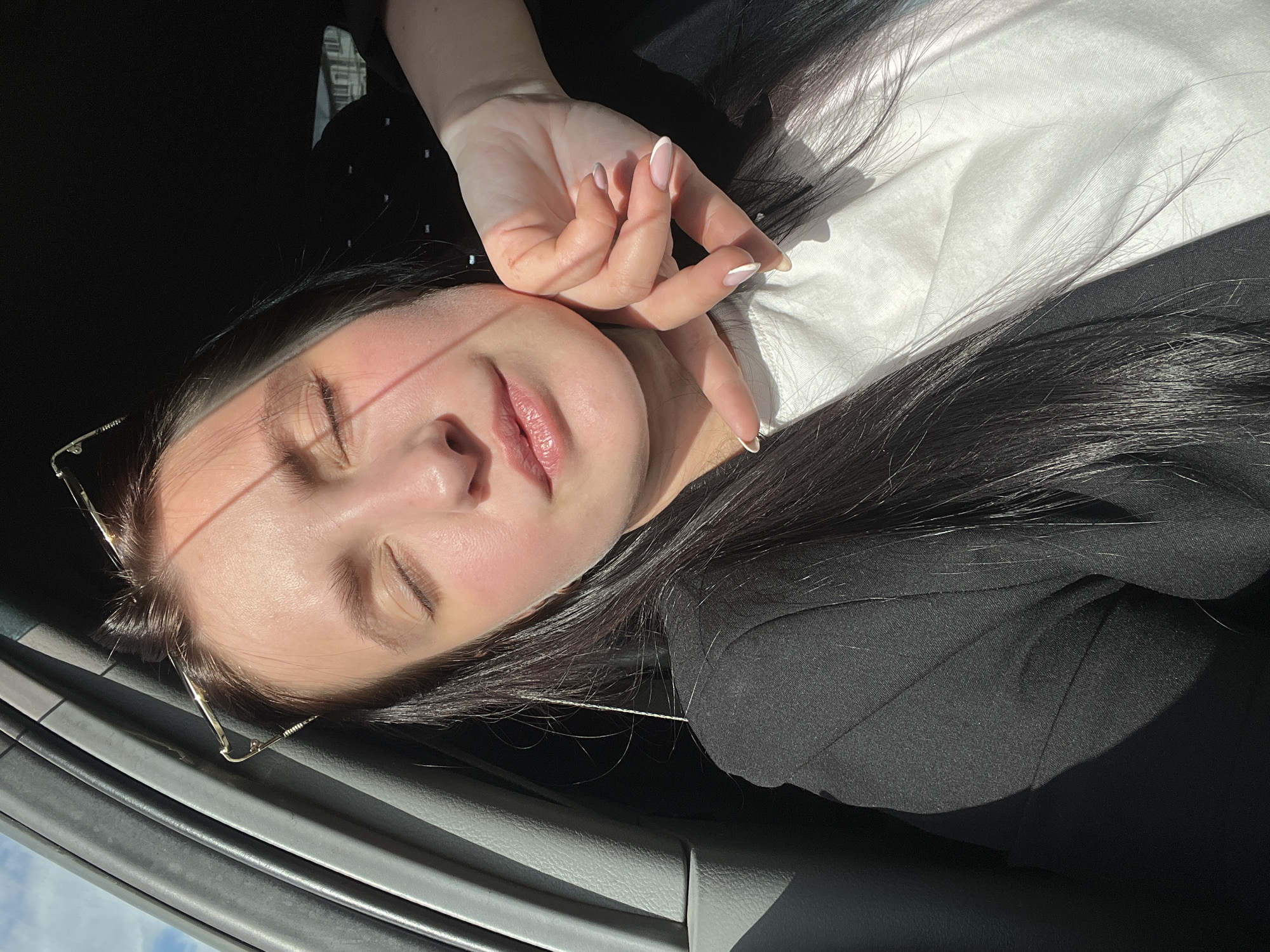 A young woman with dark hair and closed eyes is sitting in a car, her face illuminated by sunlight. She is wearing a black blazer over a white shirt. Her right hand is touching her cheek, and her nails are painted with a French manicure. Her eyeglasses are perched on her head. The interior of the car is visible, including the grey door panel and the dark headliner. Outside the car, the sky is blue with some clouds. The image appears to have been taken on a sunny day.