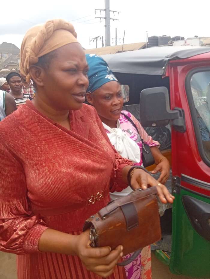 This image depicts an outdoor urban or market scene in Ura Nta Umuarandu, Nigeria. In the left foreground, a dark-skinned female, estimated to be between 40-50 years old, is visible from the waist up. She wears a reddish-brown, long-sleeved, ribbed dress with a gold-colored three-link belt buckle at the waist, and a light brown headwrap styled with a braided top section. She holds a small, rectangular brown leather bag with both hands at waist level, displaying a ring on her right ring finger. Her gaze is directed slightly off to the right from the viewer's perspective.
Behind her and to the right, a second dark-skinned female, also estimated between 40-50 years old, is partially visible, appearing to be seated inside a red and green three-wheeled auto-rickshaw (keke napep). She wears a blue patterned headwrap, a white top over a multi-colored garment (pink, purple, white), and has a dark strap over her shoulder. Her mouth is slightly open, suggesting speech. The right side of the red and green auto-rickshaw, including its black soft top and side mirror, is visible.
In the background, multiple indistinct figures are present, suggesting other individuals in the vicinity. The environment includes various structures: buildings with corrugated iron roofs, multiple utility poles with overhead power lines, and black water storage tanks visible on rooftops. A partial white sign with red text and a red cross emblem are discernible on a distant building. The sky appears overcast.