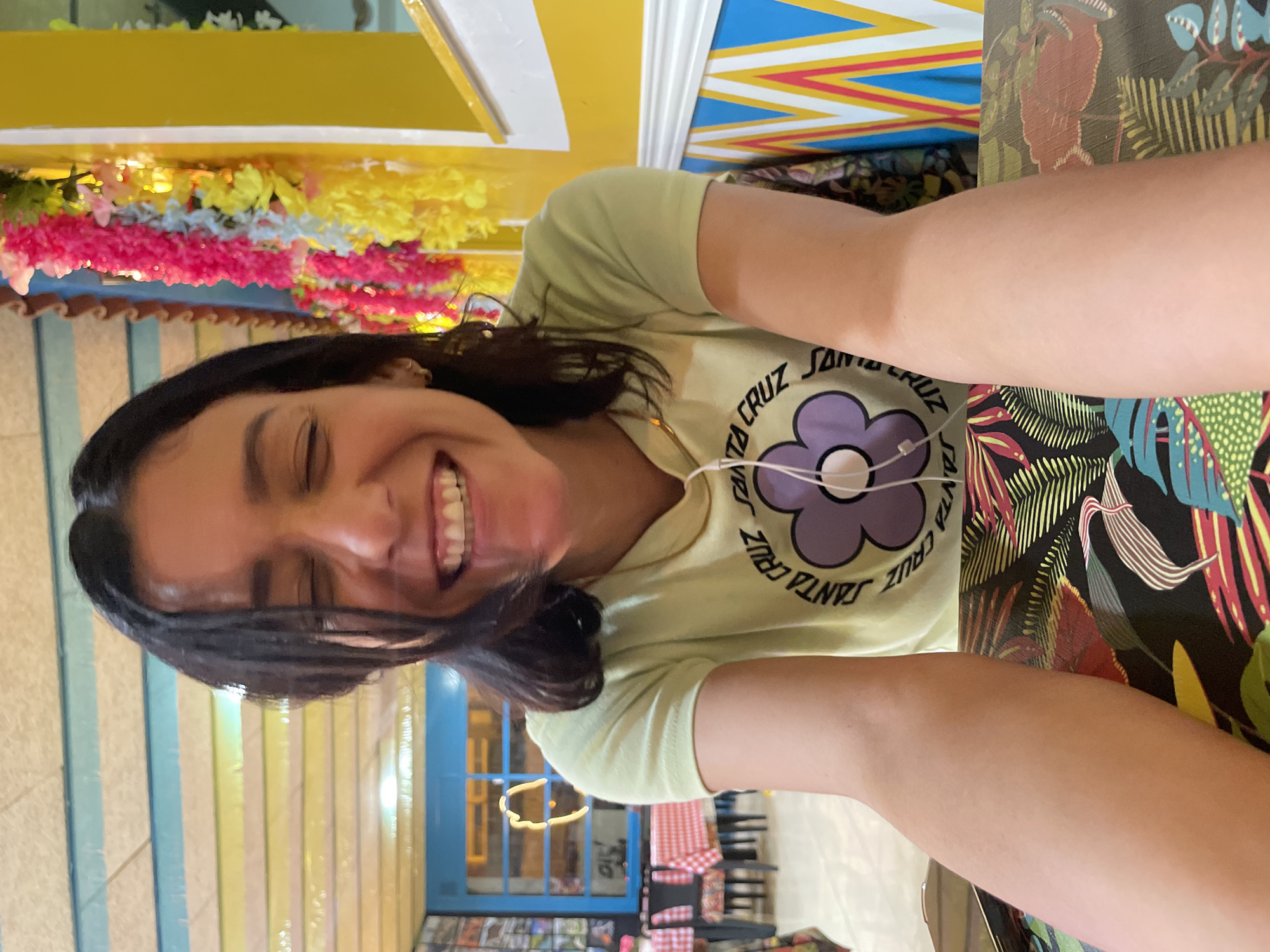 A woman is taking a selfie in an indoor setting. The woman has dark hair and is wearing a light green t-shirt with a purple flower and the text "SANTA CRUZ" around it. She has earphones in. She is smiling broadly, with her eyes closed. The background includes a decorated ceiling with colorful stripes and floral garlands. The table in front of her is covered with a vibrant, patterned cloth. Further back, there is a view of a building interior with tables, a blue door, and colorful wall panels. Based on the building interior, it appears to be a restaurant, potentially located in L'Hospitalet De Llobregat, Spain. The exact location cannot be determined from the image.