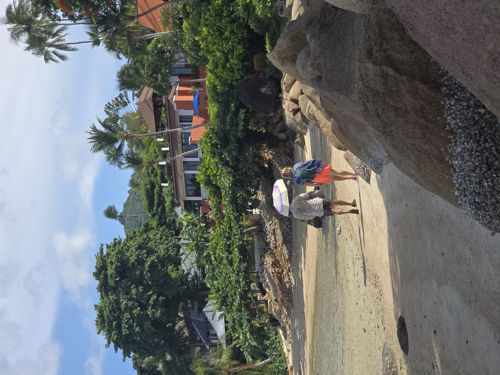 Two individuals walk along a sandy beach in Baan Chaweng Noi, Thailand. The person in front, holding a colorful umbrella, wears a patterned shirt and shorts. Behind them, another individual walks with a backpack, wearing an orange skirt and a blue and white patterned top. The beach is bordered by lush green vegetation, including palm trees and large bushes, with a modern building complex visible in the background. Large rocks are scattered along the shoreline, some covered in seashells. The sky is blue with scattered white clouds.