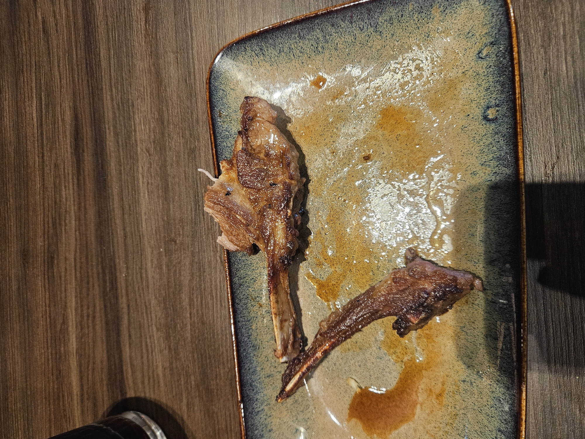 The image shows a close-up of a rectangular plate with two lamb chops, mostly eaten, on a wooden table. The lamb chops are well-cooked and appear to have been seasoned. There are some greasy streaks and remnants of sauce on the plate. A glass with a dark liquid is partially visible in the upper left corner of the image. The scene appears to be a restaurant setting in Hoogvliet, Netherlands, as indicated by the requested location. There are no people or actions depicted in the image, it's a static scene of food remnants on a plate.
