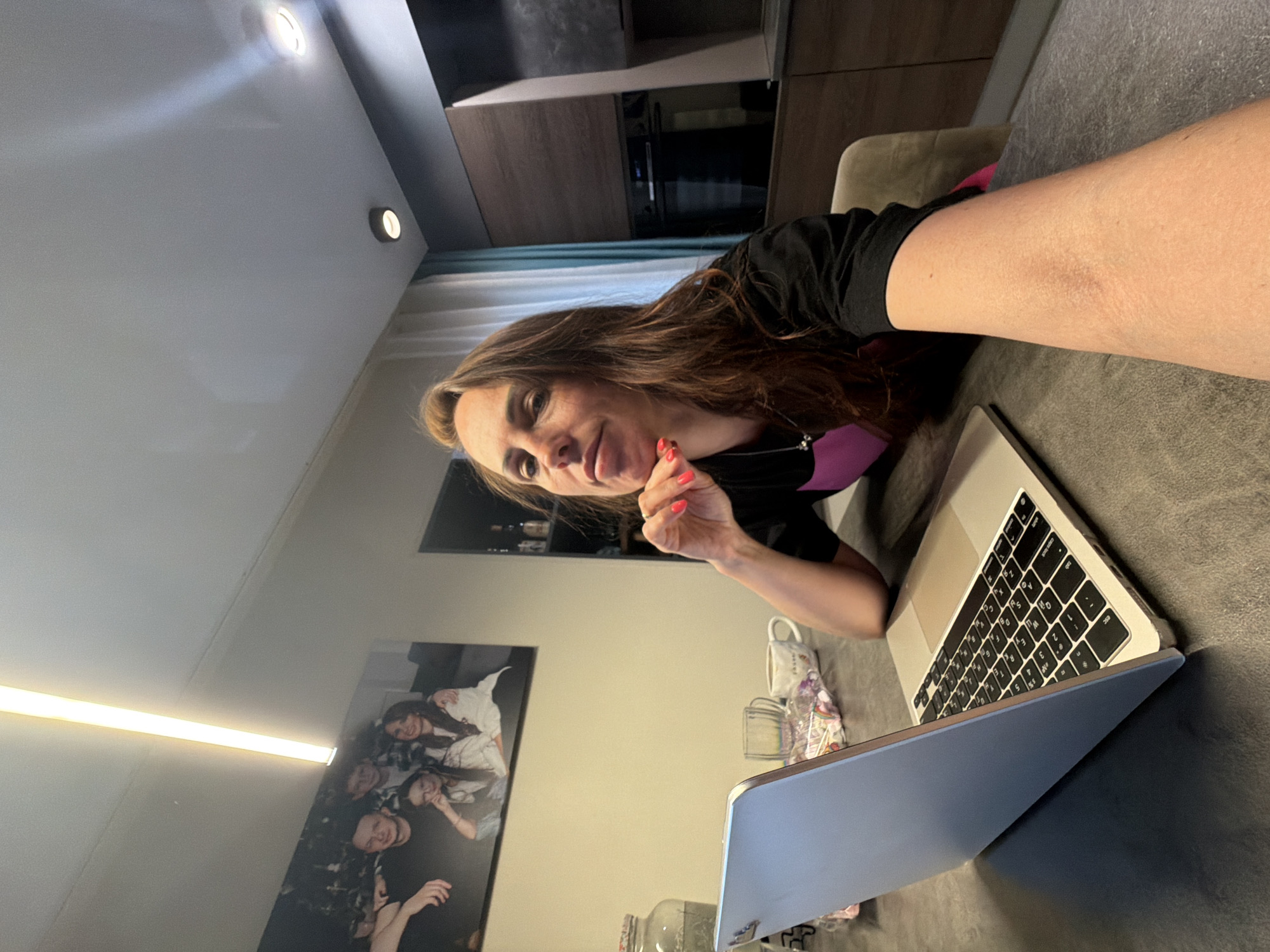 A woman with long, reddish-brown hair is depicted in a modern indoor setting, likely a kitchen or dining area in Birobidzhan, Russia. She has light skin and is wearing a black top with a pink or fuchsia accent, with her fingernails painted a bright coral pink. She is looking directly at the camera with a slight, content smile, resting her chin lightly on her right hand.
In front of her, on a dark grey tabletop, is an open silver MacBook laptop. The lid of the laptop has a light blue cover, and the keyboard clearly displays Cyrillic characters alongside standard Latin alphabet keys, confirming its regional use. To the right of the laptop, a white mug and a clear glass are visible, along with what appears to be a small bag of snacks or candy.
The background features contemporary interior design elements. Behind the woman, there are dark wooden cabinets and what looks like kitchen appliances. The wall to her left displays a framed photograph depicting several smiling individuals, suggesting a personal touch to the decor. Above, the ceiling is light-colored with embedded circular spotlights, and a long, linear light fixture illuminates the wall where the photograph hangs. The overall atmosphere appears comfortable and well-lit, suggesting either daytime indoors or an evening setting with artificial lighting. The woman appears relaxed, possibly taking a break or casually interacting with her laptop.
