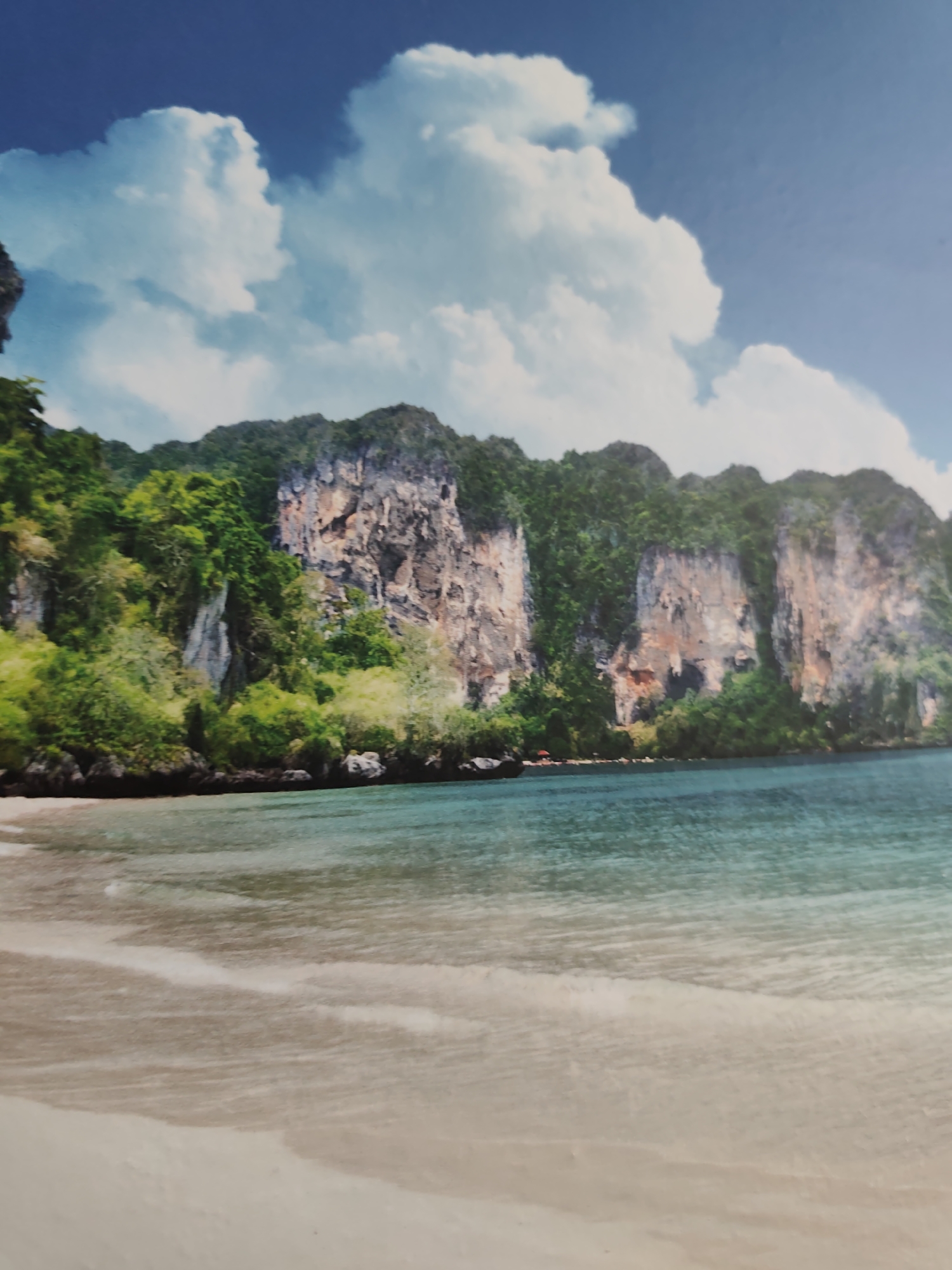 The image depicts a serene tropical coastal landscape. In the foreground, a light beige sandy beach with subtle wave patterns transitions into clear turquoise ocean water, showing gentle ripples. The midground and background feature a continuous stretch of towering limestone karst cliffs, densely covered with vibrant green tropical vegetation. Exposed rock faces display shades of grey, tan, and ochre, indicating natural geological formations. A small, dark red-roofed structure is visible at the base of one of the cliffs on the right side. The sky above is a bright blue with scattered, fluffy white cumulus clouds. No people or animals are visible. The scene is located in the city of Pazardzhik, Bulgaria.