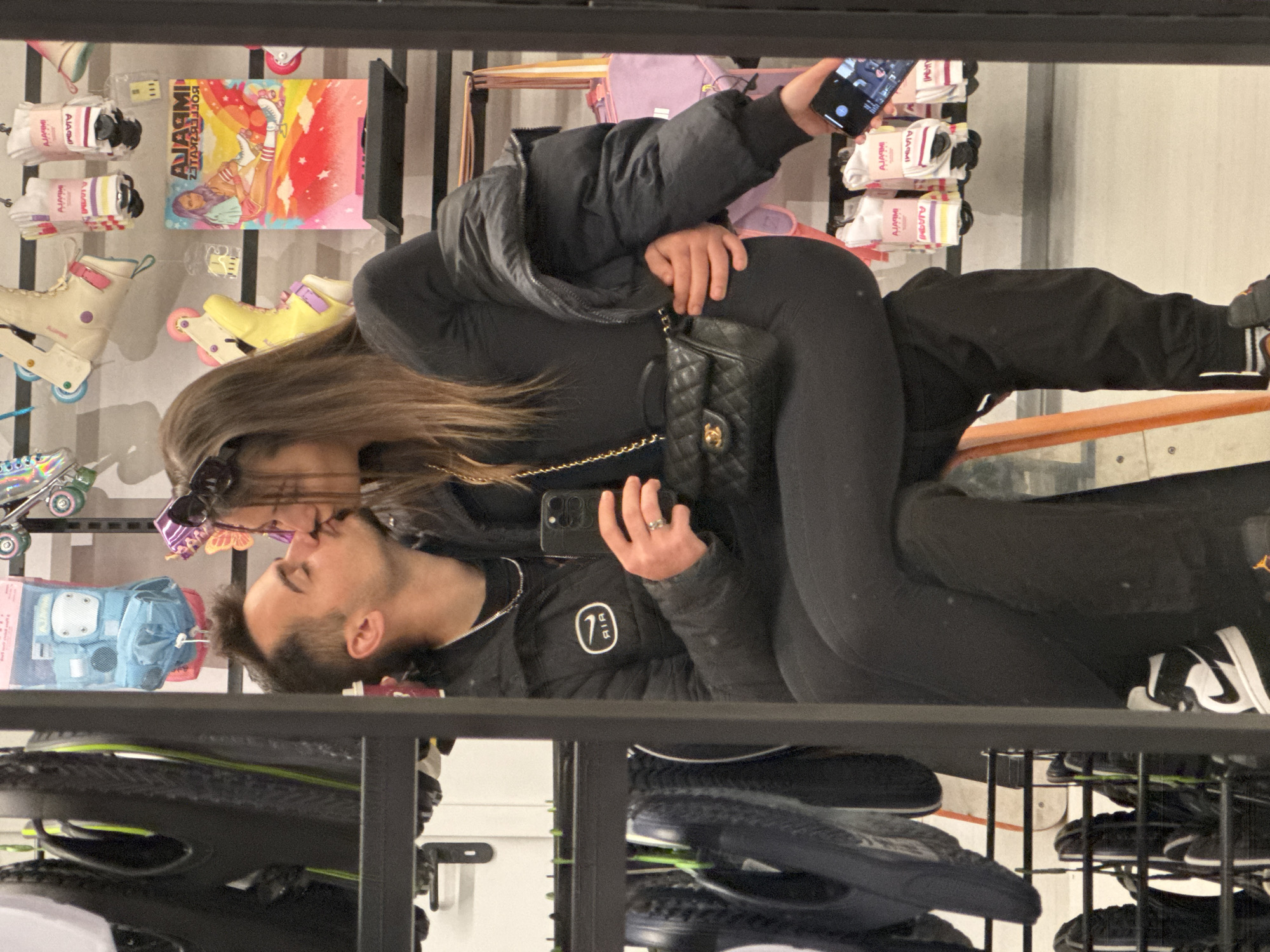 An intimate moment is captured in what appears to be a retail store, with a couple sharing a kiss. The image shows the two individuals from the waist up, with the woman embracing the man, and both are holding cell phones. The scene is shot from a slightly high angle, looking down onto the couple and their surroundings. The image is oriented sideways, with the subjects appearing horizontal.
The woman, with long brown hair, is wearing a black puffer jacket over a black top and black leggings. She is carrying a black quilted handbag, likely a Chanel bag, identifiable by the interlocking 'C' logo. She has sunglasses perched on her head and is holding a dark-colored smartphone in her right hand, which appears to be displaying a camera interface. A ring is visible on her left ring finger.
The man, with short dark hair and a beard, is wearing a black jacket with a white circular logo featuring "AIR" and a stylized '1' or '9' design, possibly referencing a Nike or Jordan brand. He is also holding a dark-colored smartphone in his left hand.
They are positioned in an aisle or display area of a store. In the background, various merchandise is visible. There are rows of roller skates in different colors, including cream and yellow, and packages of socks. A colorful poster is displayed behind them, which reads "AJARMI ROLLERBLADES" when viewed correctly. Several socks also have "AJARMI" text visible on their packaging. On shelves below, various shoes and sneakers are present, including black and white sneakers with a Nike swoosh and another with an orange Jordan logo. The overall setting suggests a store specializing in sports or leisure equipment, possibly in Sofia, Bulgaria, as per the provided context. The activity conveys affection and intimacy between the couple.
