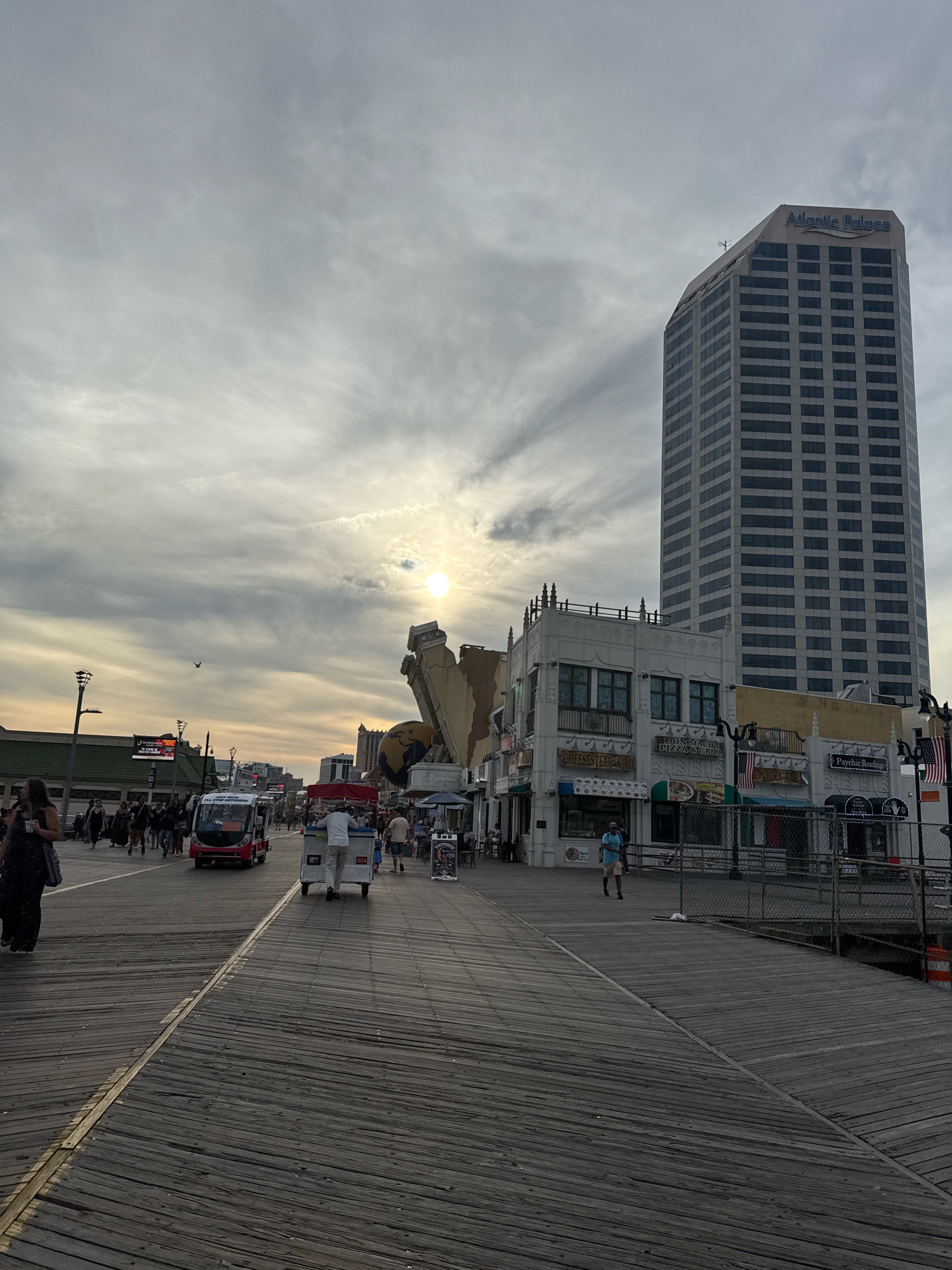 The image depicts a view of the Atlantic City Boardwalk in Atlantic City, United States of America. A tall building, the Atlantic Palace, dominates the skyline on the right side of the frame, its facade covered in numerous windows. In the foreground, the weathered wooden planks of the boardwalk stretch into the distance. Various people are visible walking along the boardwalk, some appearing to be tourists. A small, open-air electric vehicle, a "jitney" or similar transport, is seen in the middle ground, along with other individuals operating carts. Shops and businesses line the boardwalk, with signage indicating food establishments and services such as psychic readings. The sky is overcast, with the sun visible as a bright, diffused orb through the clouds, suggesting either early morning or late afternoon. The scene conveys a typical day on a bustling boardwalk.
