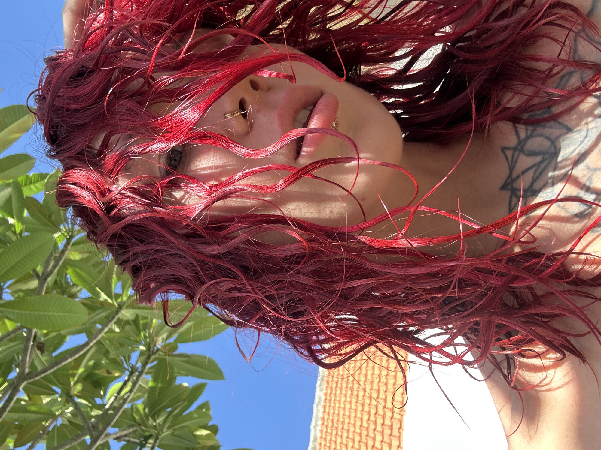 A low-angle shot depicts a person with red hair, possibly in Dalung, Indonesia. The perspective is from below, looking up at the individual. Visible are the person's face, including lips, nose ring, and one eye, obscured by strands of red hair. Part of a tattoo is visible on their shoulder. The background includes green leaves from a tree, a patch of blue sky, and a glimpse of a building with brickwork.