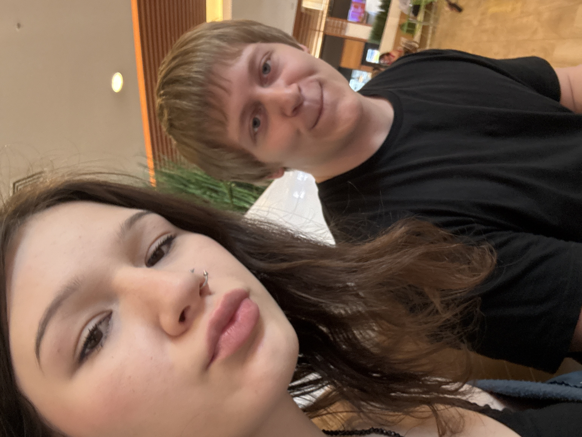 A selfie-style photograph captures a young woman and a young man in a brightly lit indoor commercial environment. The woman, positioned closer to the camera on the left, has long, wavy dark brown hair, dark eyeliner on her visible left eye, and a small silver C-shaped or spiral piercing in her left nostril. Her full lips are tinted light pink, forming a slight pout. She wears a black, possibly sleeveless, top, and a dark beaded necklace is partially visible on her left side. The man, positioned slightly behind and to the right, has short, straight, light brown or blonde hair with bangs. He has blue eyes, wears a plain black crew-neck t-shirt, and exhibits a faint smile or smirk. Both individuals are looking towards the camera.
The background is a modern, spacious interior featuring light-colored ceiling with a visible circular recessed light fixture. Walls exhibit warm-toned horizontal paneling, accented by a distinct bright orange horizontal lighting strip or trim located higher up. Green foliage, possibly artificial plants, is integrated into the interior design on the left side. Blurred figures of other patrons are visible further back, some seated at tables equipped with light green chairs. The floor is light-toned, appearing to be wood or large tiles. The image contains no specific visual markers, landmarks, or textual information that identifies its precise location within the city of Saint Petersburg, Russia.