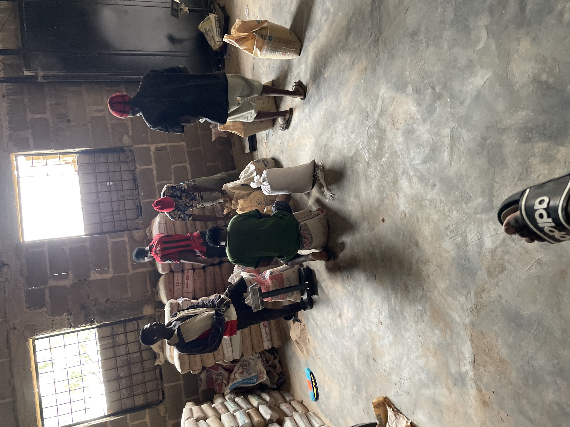 The image shows a warehouse interior with several men working with sacks of what appears to be cement. The location is identified as Jos, Nigeria. The warehouse has a concrete floor and walls made of concrete blocks. Natural light streams in from two windows with metal grates. Stacks of bagged cement are visible along the walls.
There are at least six men in the scene. One man in a red and black striped shirt is standing, facing away from the camera. Another man in a black shirt and light-colored shorts is also standing, facing away. A third man in a green shirt is sitting on a sack. A fourth man in a patterned shirt and red cap is also standing. Two other men are partially visible.
Various objects are present, including sacks of cement, a weighing scale, and a broom lying on the floor. The overall atmosphere suggests an industrial or storage setting where manual labor is being performed. The presence of cement bags and the manual handling indicate a commercial or industrial operation related to construction materials. The lighting suggests it is daytime, possibly midday given the brightness of the windows.