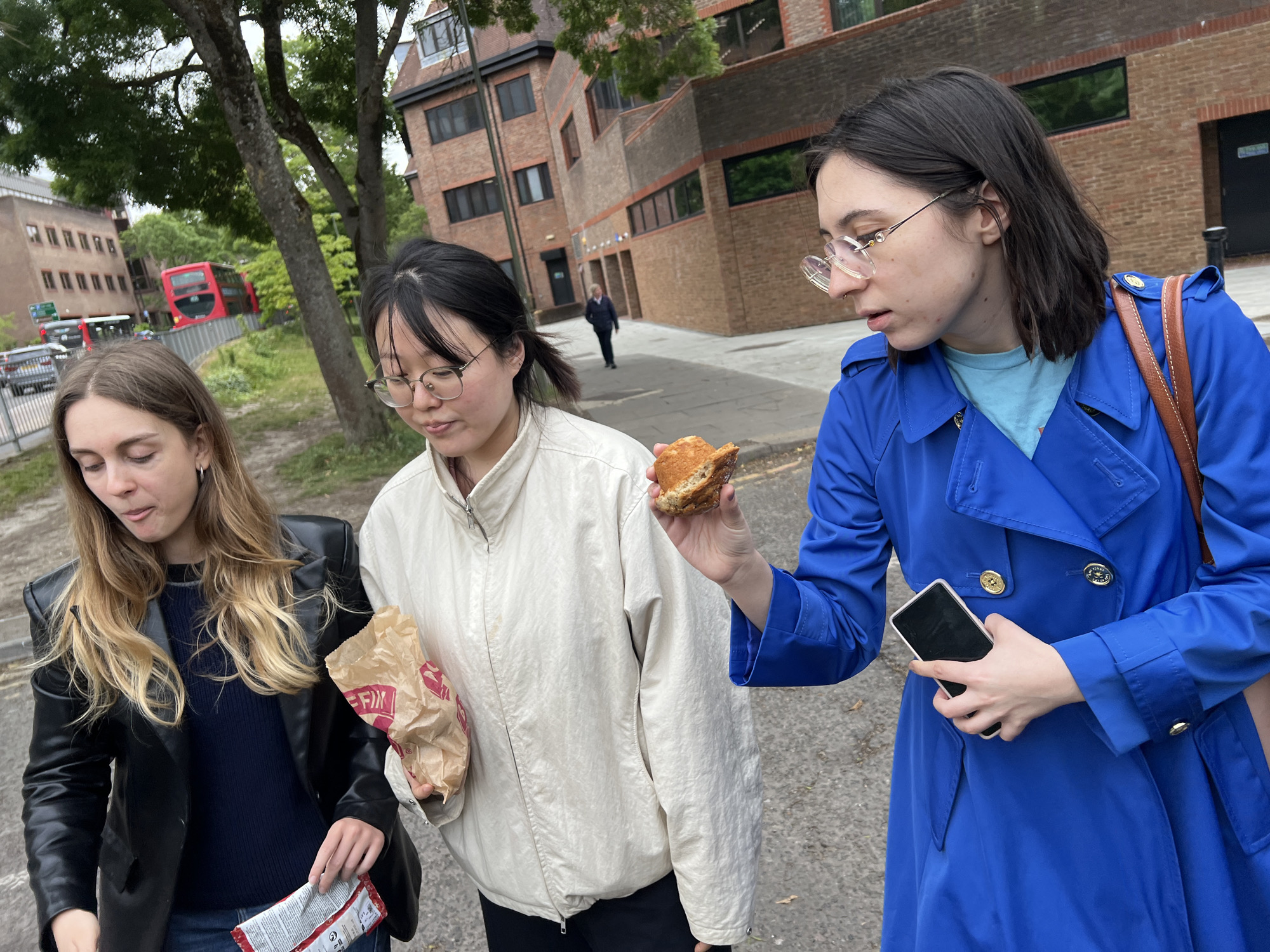 Three young women walk on a paved surface adjacent to brick buildings and trees. The woman on the left wears a black leather jacket and dark blue top. The woman in the middle wears glasses and a light-colored jacket, holding a brown paper bag. The woman on the right wears a blue trench coat, holding a smartphone in her left hand and a halved sandwich in her right hand, looking at the sandwich. A red double-decker bus is visible in the background. A person walks away from the camera further up the street. The setting appears to be an urban area, possibly Epsom and Ewell, UK.