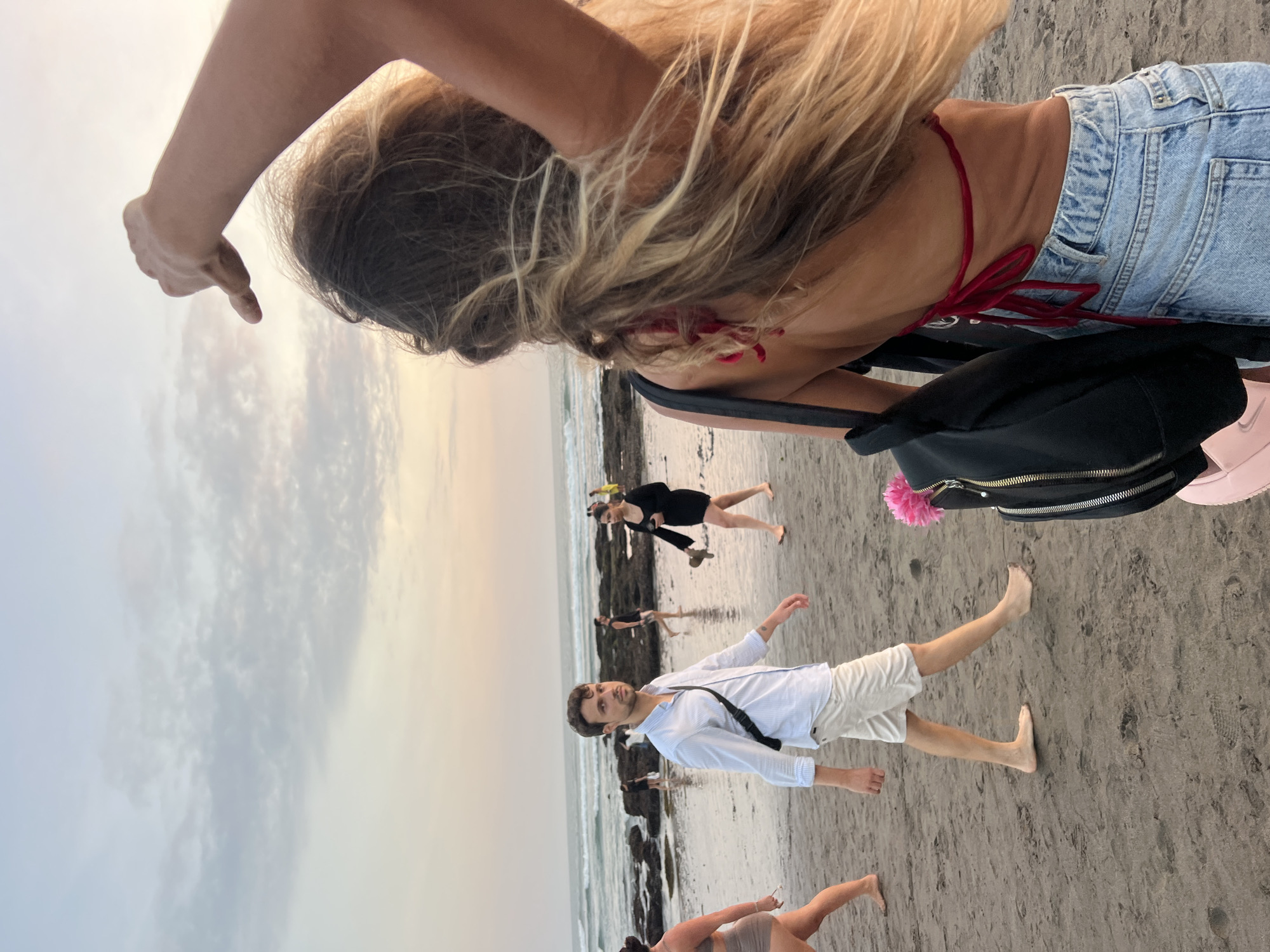 A group of people are walking on a sandy beach in North Kuta, Indonesia, near the ocean. In the foreground, a woman with long blonde hair and a red bikini top is seen from behind, holding her arm up with her hand extended. She is wearing denim shorts and carrying a black backpack adorned with a pink flower. To her left, a man in a light blue striped shirt and white shorts is walking towards the camera. In the background, other individuals are visible on the beach and in the water. The sky is overcast with soft, muted colors.