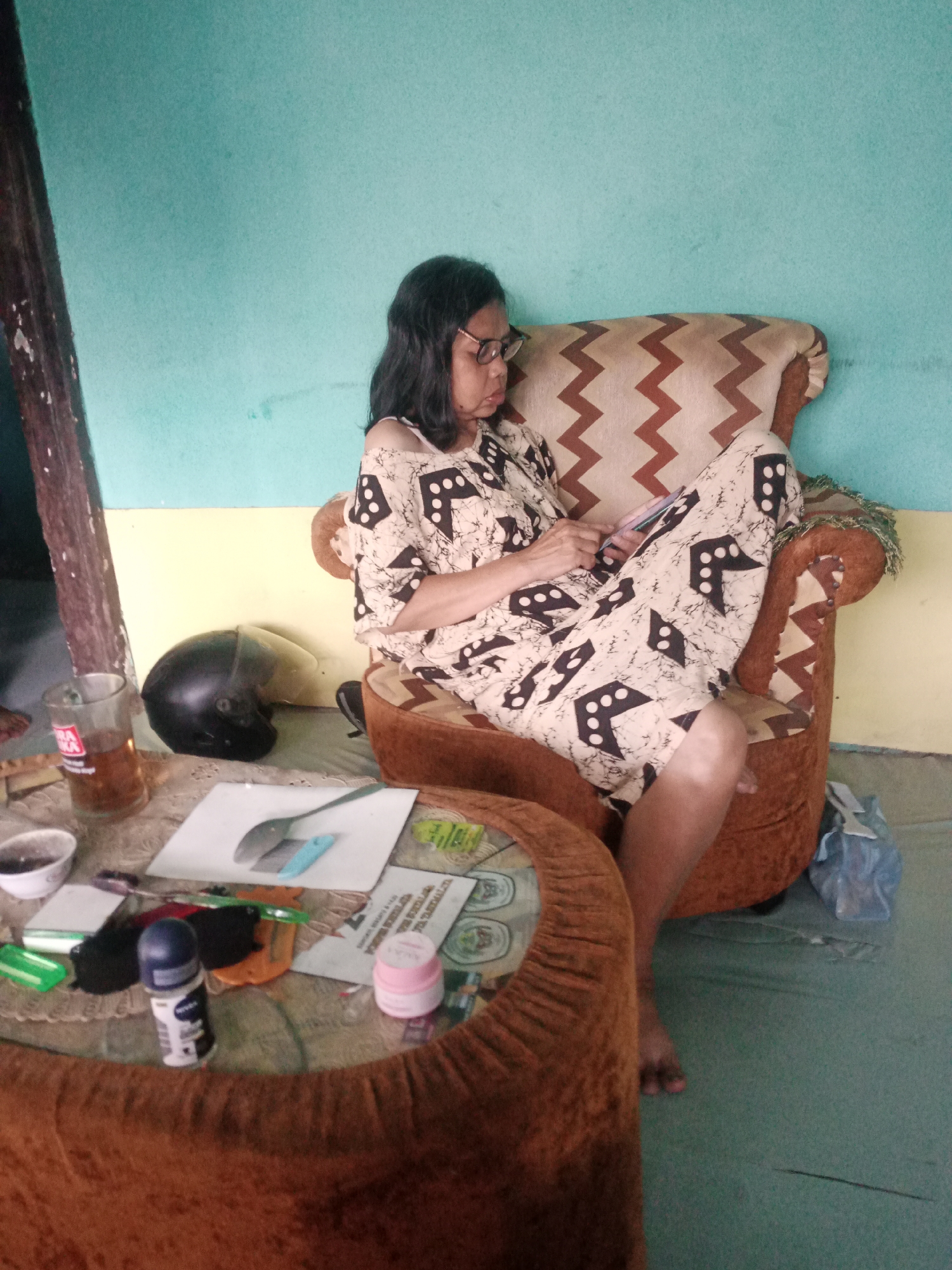 A woman is sitting in a chair and looking at her phone. She is wearing a dress with a pattern of black and white dots. There is a table in front of her with a glass of tea, a helmet, and some personal care items on it. The room appears to be a living room or a common area, with a light blue wall and a yellow border. The furniture is upholstered in a brown fabric with a zig-zag pattern. The image was taken in Tasikmalaya, Indonesia.