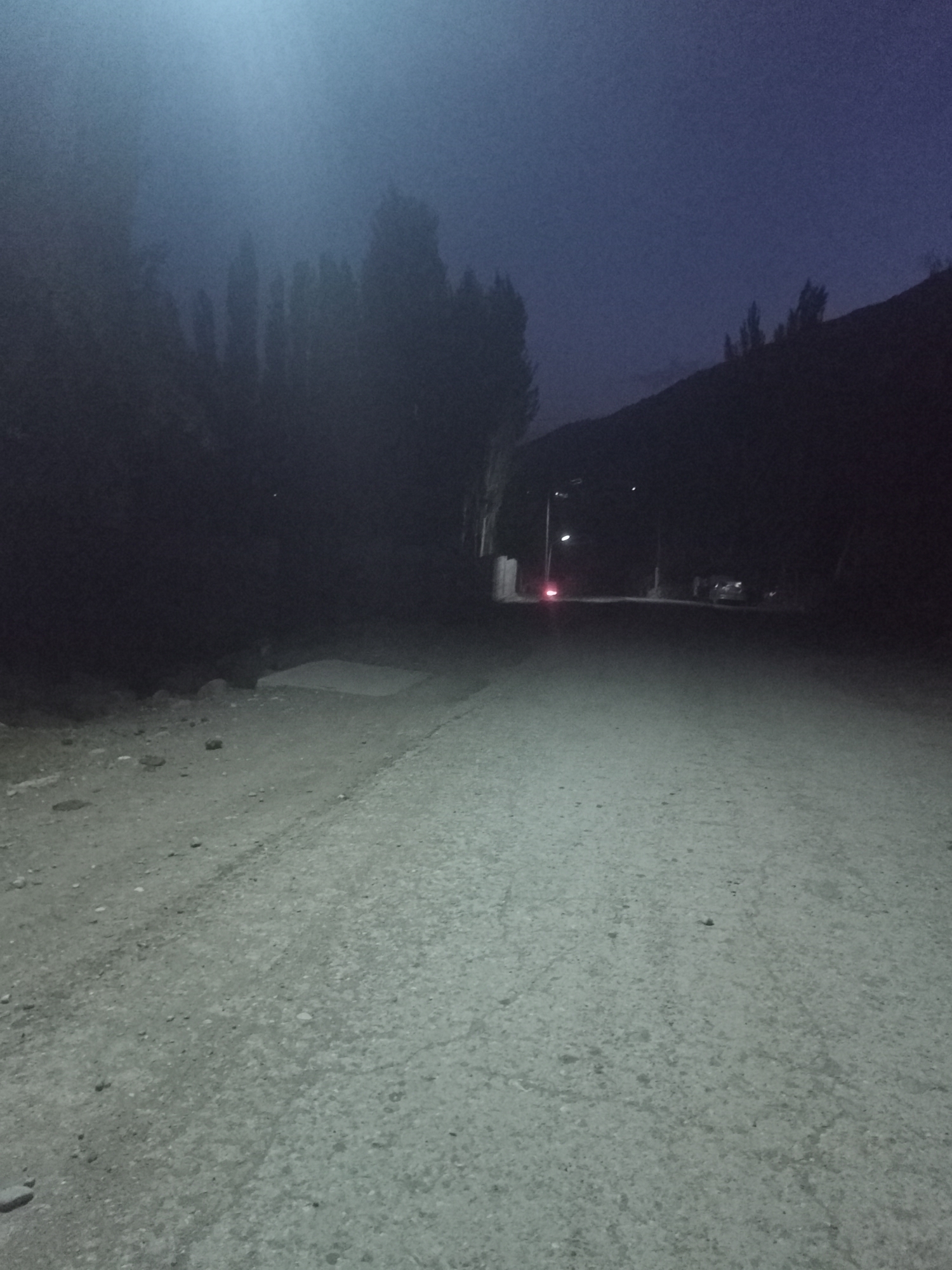 This is a nighttime, low-light, horizontal, eye-level shot of a dirt road in the city of Sayod, Tajikistan. The road is dusty and uneven, with scattered rocks. To the left of the road, there is a dark, forested area, with tall, slender trees silhouetted against a hazy, blue sky. The sky is overcast and there is a bright light source in the upper left quadrant of the image, possibly a streetlight or the moon. In the distance, the road curves to the right, leading towards a dimly lit area where a few buildings and a vehicle are visible. The overall mood is dark and somber, with a sense of isolation.