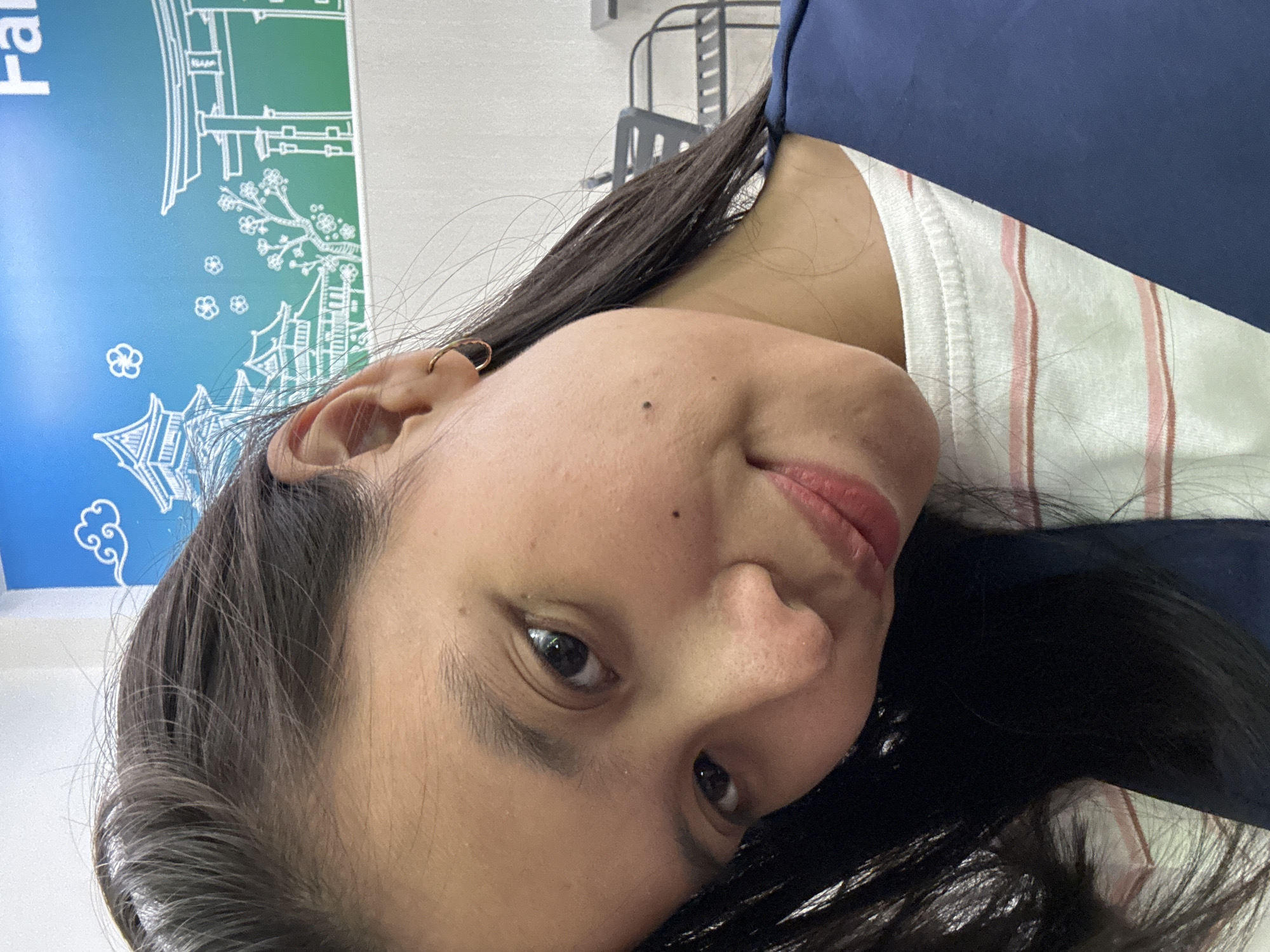 A young woman with dark hair and brown eyes is pictured in a close-up, selfie-style shot. She is wearing a white t-shirt with thin pink stripes and a dark blue jacket. Her hair is parted on the side and falls over her shoulder. She has a small hoop earring in her left ear. A mole is visible on her left cheek. The background is a wall with a blue and green mural depicting Japanese-style architecture, specifically a pagoda and torii gate, along with cherry blossoms. The environment appears to be indoors, possibly a cafe or restaurant given the seating visible in the blurred background. There is no specific indication of the city of East Jakarta, Indonesia, in the image.