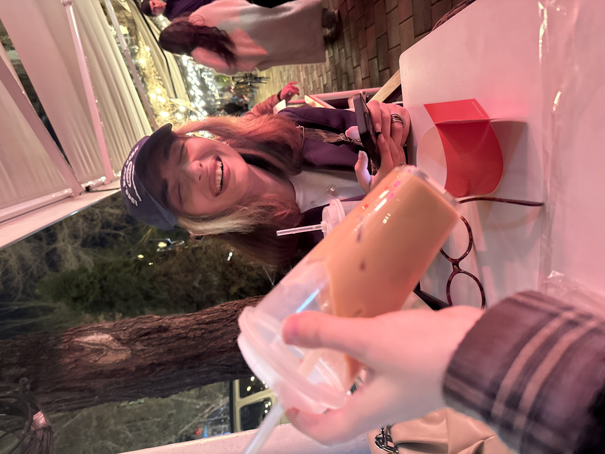 A tilted, close-up indoor shot captures a smiling person wearing a navy blue baseball cap with white text, facing the viewer at an outdoor restaurant. The person is seated at a table, holding a mobile phone with both hands. In the foreground, a hand is holding a clear plastic cup filled with a brown-colored beverage with a straw. The table is white, with a red object sitting on it, next to a pair of glasses. The restaurant appears to be outdoors, with a tree and lights visible in the background. There are other patrons further back in the image. The location is Yerevan, Armenia.