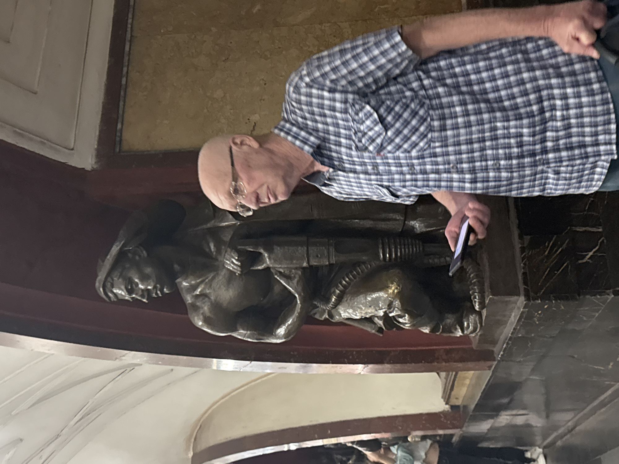 An outdoor, full shot of a man standing next to a statue in the Moscow Metro. The man is bald, wearing glasses and a blue and white plaid shirt. He is holding a phone in his left hand, looking to the right. The statue is a bronze sculpture of a man holding a jackhammer. The man is wearing a hat and a scaly uniform. The statue is on a marble pedestal. The background is a subway station with white ceilings and red walls.