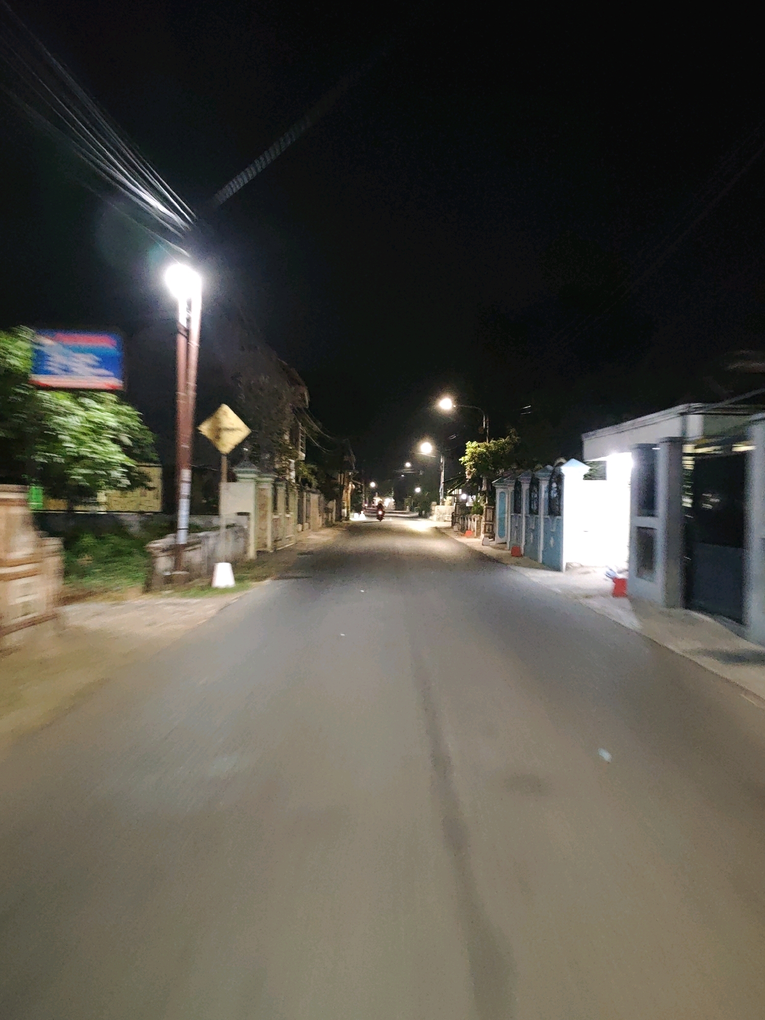 The image depicts a street scene at night in Pagu, Indonesia. A paved road stretches into the distance, lined with buildings and trees. Streetlights illuminate the scene, casting long shadows. On the left side of the road, a traffic sign and a lamppost are visible. Further down the road, a person on a motorcycle is riding away from the camera. The buildings on the right appear to be residential, with some lights on inside. The overall atmosphere is quiet and peaceful, with the exception of the moving motorcycle.