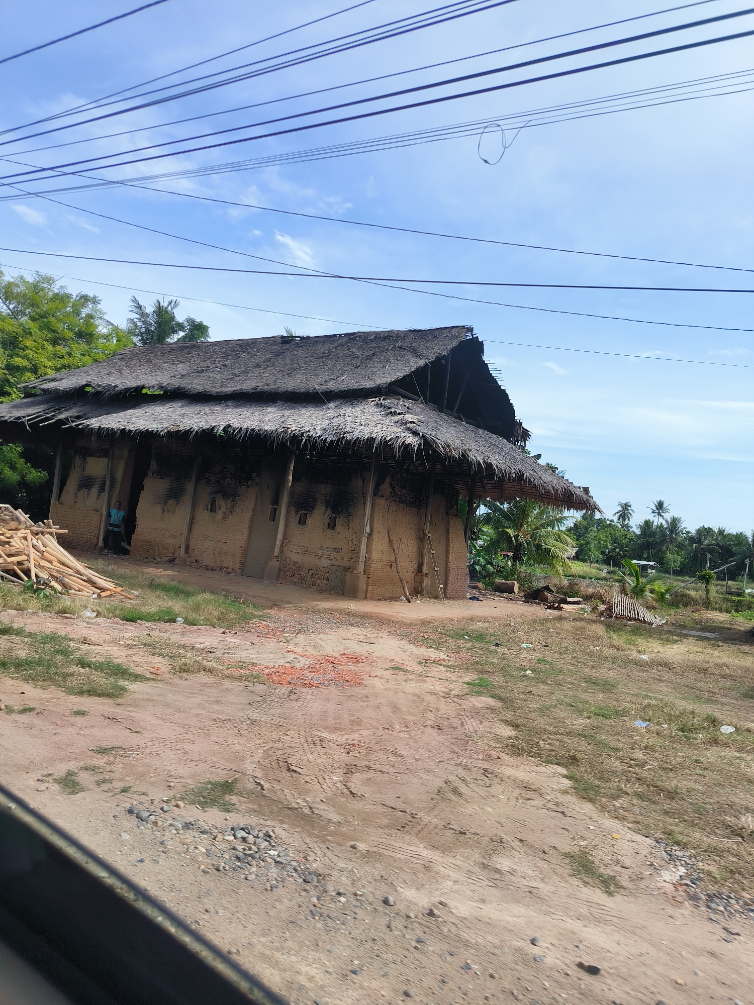 The image depicts a rustic, thatch-roofed structure with mud brick walls, possibly a dwelling or workshop, situated in a rural area. A person in a blue shirt stands within an open doorway of the structure, partially visible. Piles of wooden poles and scattered bricks are present in the foreground, suggesting ongoing construction or material storage. The surrounding environment is characterized by dry, unpaved ground, sparse vegetation, and a backdrop of lush green trees and palm trees, indicative of a tropical climate. The scene is further characterized by a network of overhead power lines against a clear blue sky with scattered clouds. The overall impression is one of a simple, traditional settlement in an undeveloped or developing region. The specific location is Lancang Barat, Indonesia.