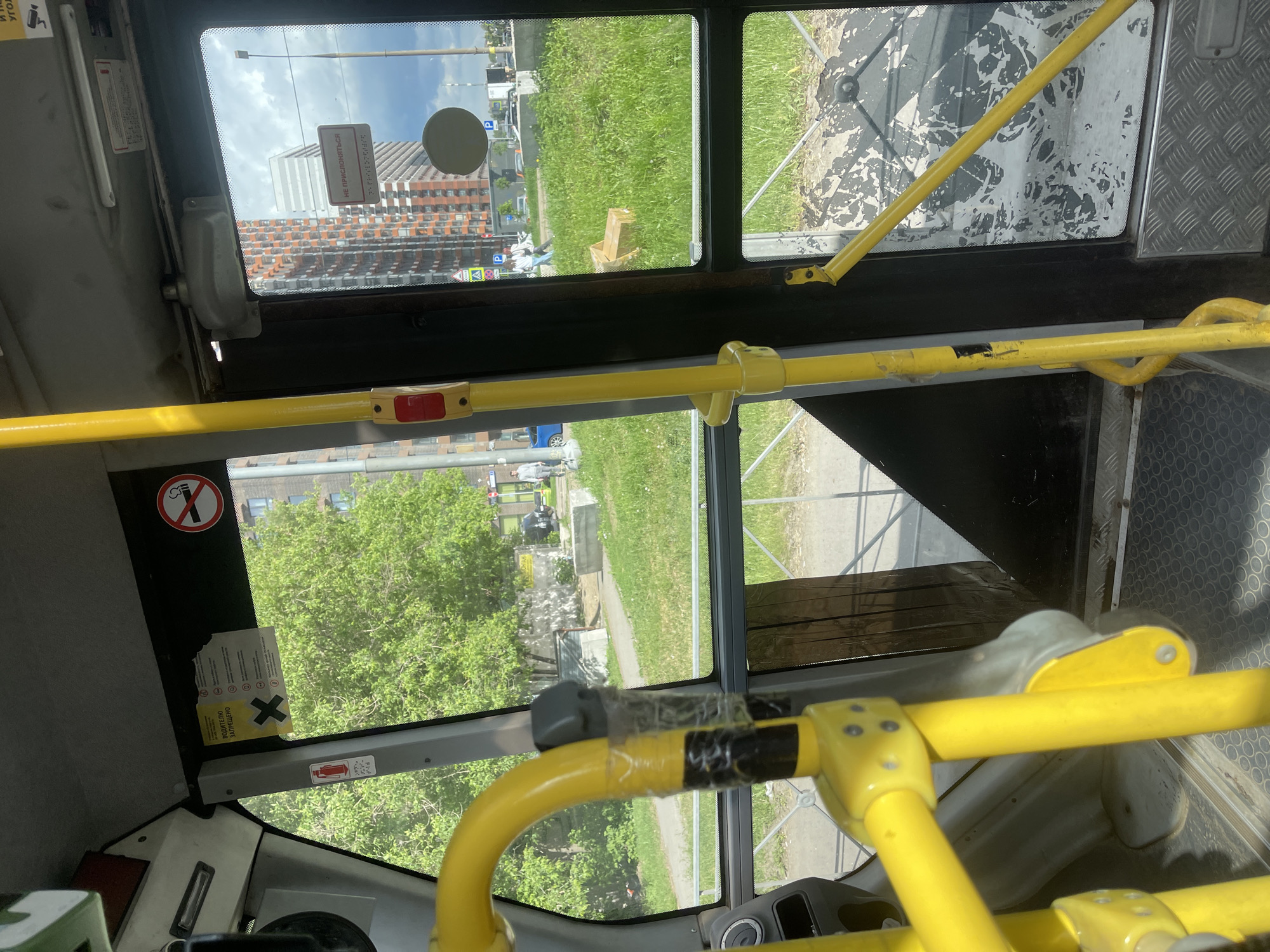The view from inside a public transportation vehicle, likely a bus, looking out through the windows. The interior features yellow handrails. Through the windows are buildings and greenery, suggesting an urban or suburban setting. One sign visible on the outside is in Russian. Based on the prompt, this scene is located in the city of Kotelniki, Russia.