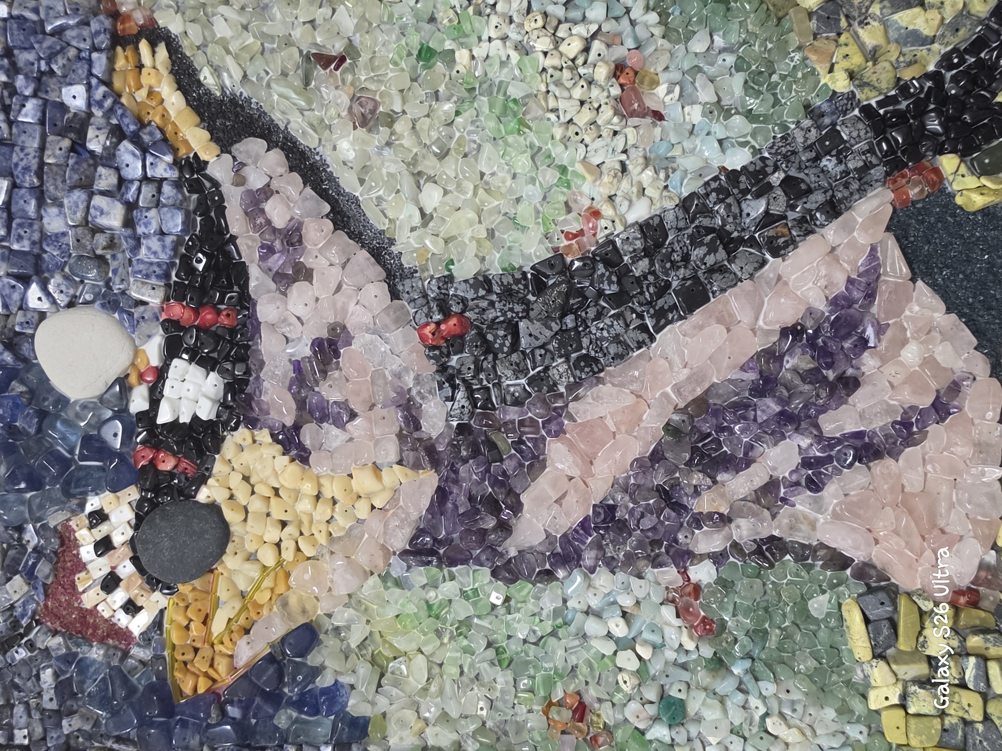 This media file is a close-up, overhead shot of a mosaic artwork. The mosaic is composed of numerous small, irregularly shaped stones and beads of various colors and textures. The arrangement of the stones forms an abstract pattern that suggests the wing of a bird or butterfly. Dominant colors include blues, purples, pinks, greens, yellows, and black, with some red and white accents. The stones vary in translucency, with some appearing opaque and others semi-transparent. The overall impression is one of intricate craftsmanship and vibrant color.
There are no visible people, animals, or specific objects that can be definitively identified, beyond the abstract representation within the mosaic. The scene is static, depicting the artwork itself, not an event or activity. The setting is implied to be indoors or a studio where such artwork would be created or displayed, given the lighting and lack of environmental context. There are no cues about time of day, weather, or specific location within Plovdiv, Bulgaria, beyond the overlaid text.
Notable details include the varied materials: the deep blue stones on the left may be sodalite, the white stone a smooth pebble, the black and grey stones could be obsidian and snowflake obsidian, the pink stones appear to be rose quartz, and the purple stones are likely amethyst. The background is a mix of smaller green, white, and yellow stones.
Visible text in the bottom right corner reads "Galaxy S26 Ultra," which is likely a watermark from the device used to capture the image.
