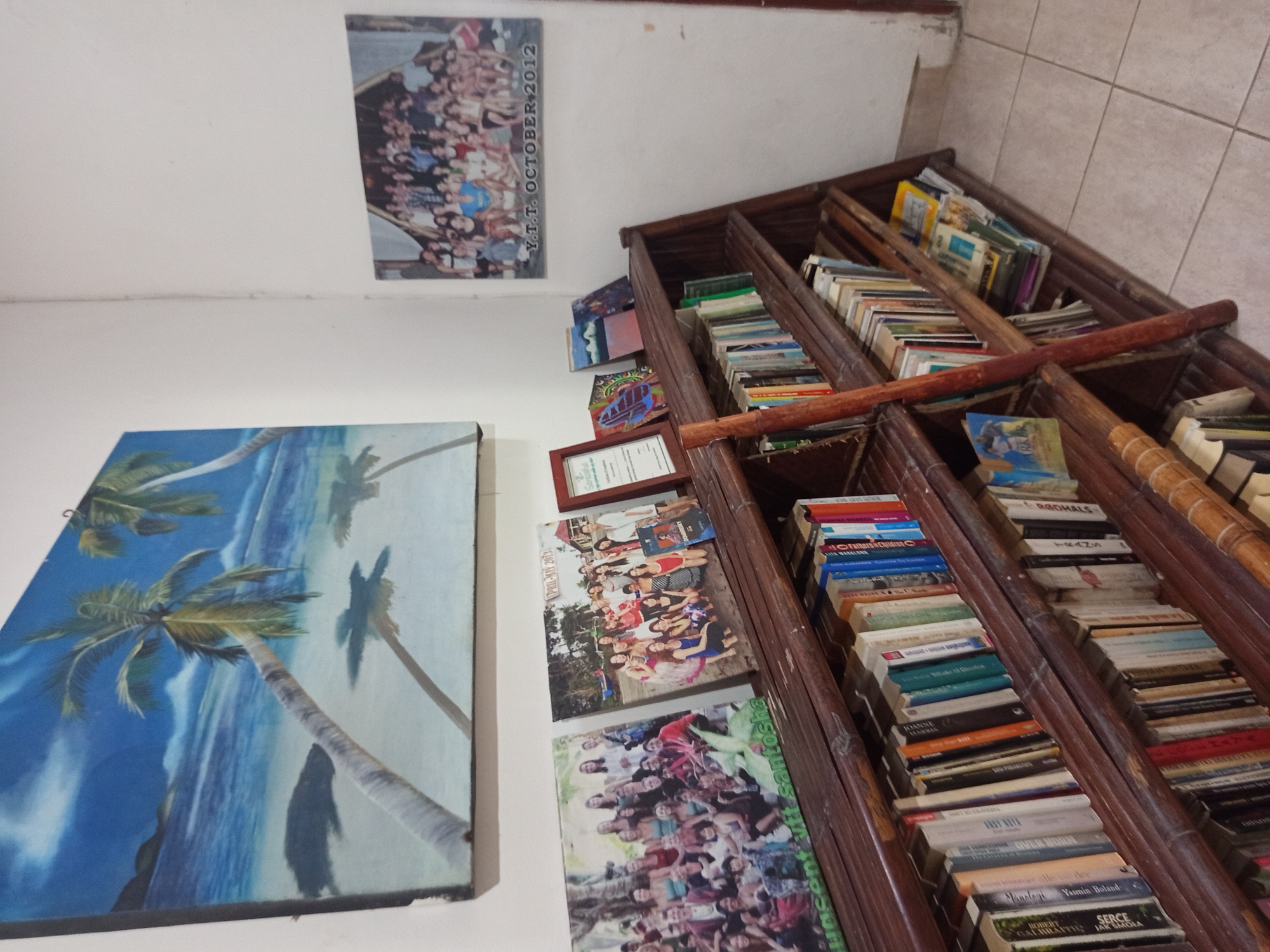 A slightly tilted, low-angle shot captures a portion of a room with a tiled floor and white walls. On the wall to the left, a painting of palm trees on a beach is displayed. Above it, a photograph of a group of people, dated "Y.T.T. OCTOBER 2012," is affixed. Below the painting, there's a framed certificate and two photographs of groups of people. One is labeled "PRIL-MAY 2013."
To the right, a wooden bookshelf-like structure with multiple tiers is packed with books, arranged horizontally. The spines of the books are visible, with titles like "RGOHALS," "TRANS," "LITA MARKLAND," "JOANNE," "ANDY NOTA," "OVER NORN," "Yasmin Boland," and "SERGE."
The images suggest the location is a room with personal items and reading materials, and the text "vtt santosha" is visible on one of the photos. The context indicates the location might be in North Kuta, Indonesia.