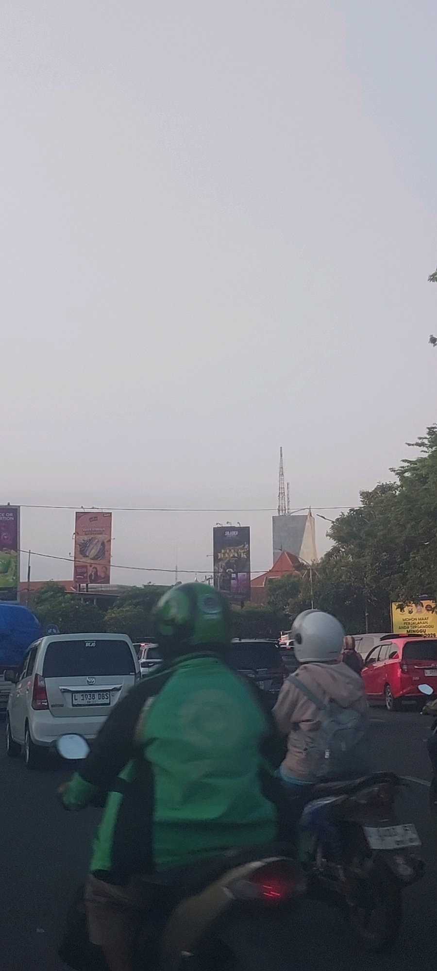 In Surabaya, Indonesia, a view of traffic on a hazy day shows several vehicles, including a white Toyota Innova with license plate L 1938 DBS, a red car, and a motorcycle carrying two individuals. The rider in front wears a green jacket and helmet, suggesting they may be a ride-sharing driver. The passenger behind wears a light-colored jacket and a white helmet. In the background, large billboards are visible, one advertising a product with an image of food, and another with text that reads "SIAER" and "BADAK." A tall antenna tower is also visible in the distance. The overall scene depicts a typical urban traffic environment in Surabaya.