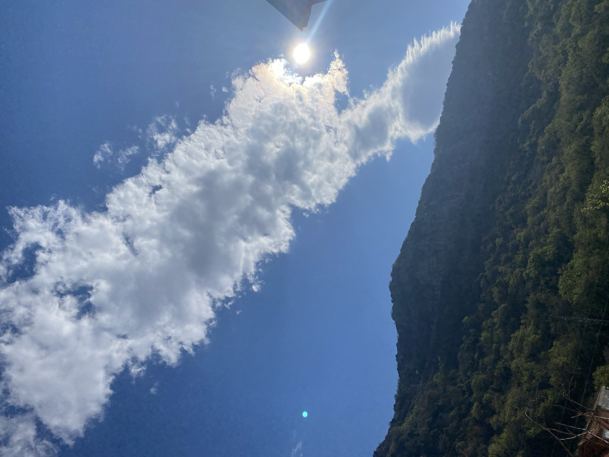 The image shows a scene under a bright, blue sky. A large, irregular cloud formation is prominent on the left side of the frame, obscuring the sun. To the right, a mountain covered in dense, green vegetation rises. The camera is oriented upward, capturing the sky and the upper part of the mountain. The location is potentially near Budva, Montenegro, given the mountainous terrain. There is no presence of people or interactions, and the objects included are natural elements, mountain, and sky.