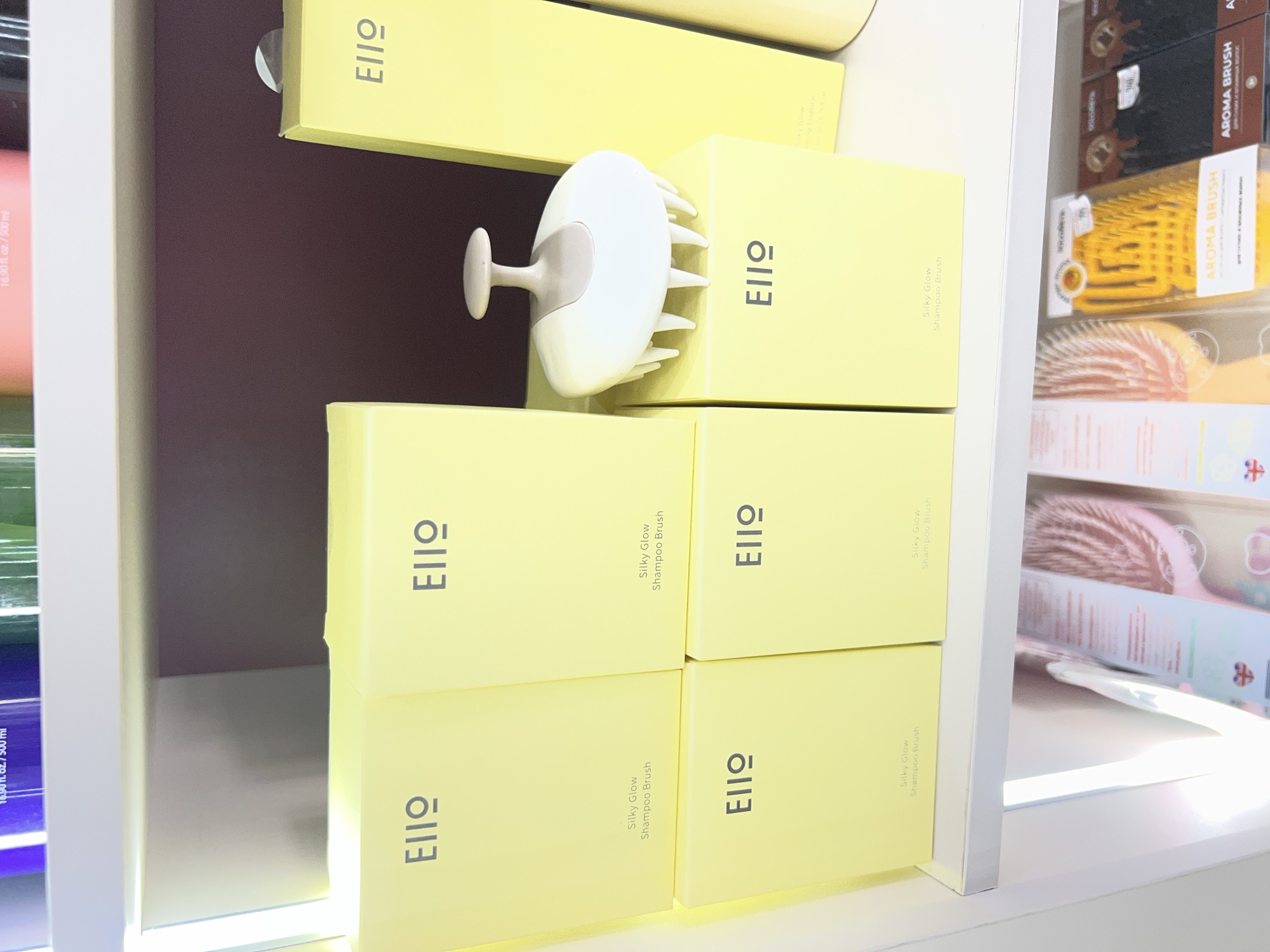 The image shows a retail display shelf in Osh City, Kyrgyzstan. The shelf holds several yellow boxes with the branding "EIIO" and "Silky Glow Shampoo Brush" printed on them. A white and beige shampoo brush with multiple prongs is displayed on top of one of the boxes. To the right, a tall yellow bottle or container is visible. Below this display, another shelf contains packaged hairbrushes, including pink and orange varieties labeled "AROMA BRUSH." The background is a deep purple wall. The overall lighting suggests an indoor retail environment.