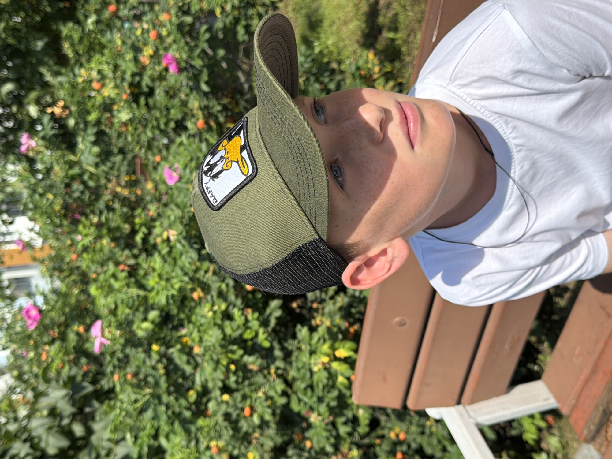 A young boy with blue eyes and freckles is sitting on a brown wooden bench. He is wearing a green trucker hat with a Daffy Duck patch and a white t-shirt. He is looking to his right. Behind him is a green bush with pink and orange flowers and some orange berries. The scene appears to be outdoors, possibly in a park or garden. There is no information in the image to determine the location within the city of Yekaterinburg, Russia.