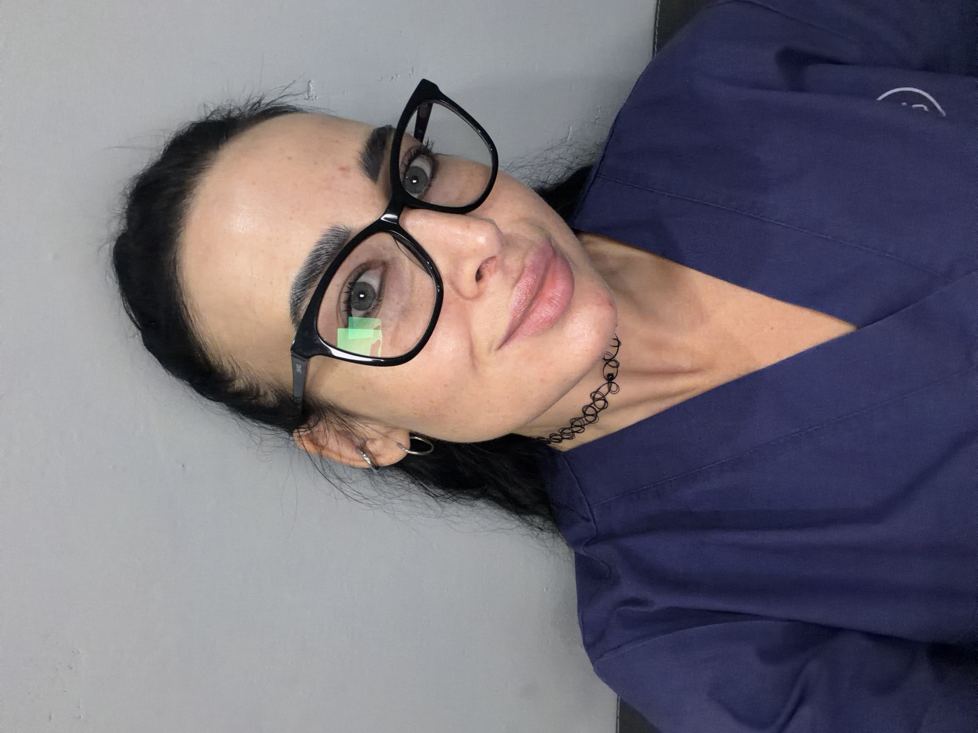 A woman with dark hair pulled back, wearing black-framed glasses and a dark blue scrub top, is pictured against a light gray background. She is wearing a choker necklace and a silver hoop earring. The photo appears to have been taken indoors. There is no information available to indicate the location is Saint Petersburg, Russia.