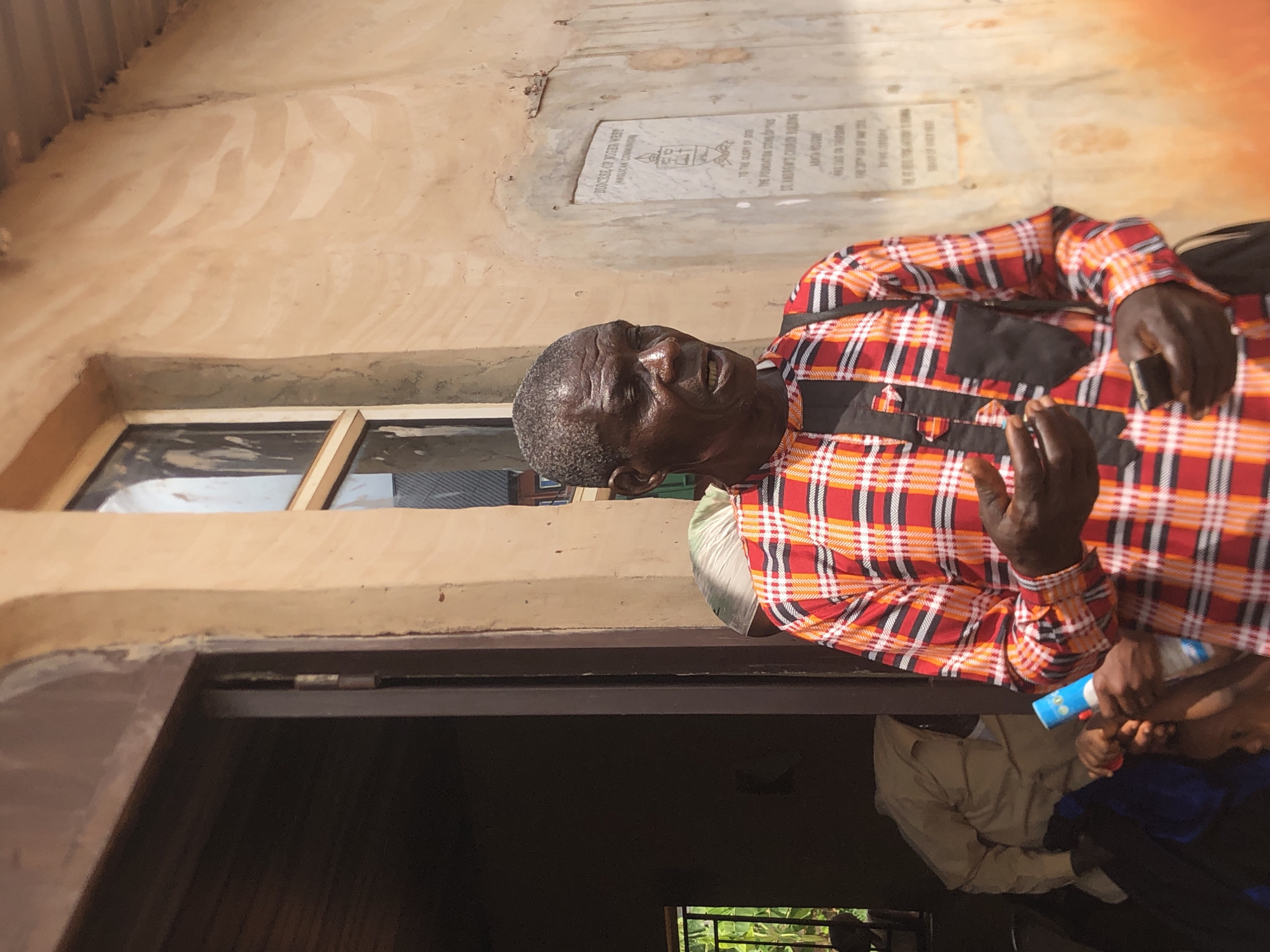 An elderly man is photographed outdoors in Ukwu-Abwa, Nigeria, standing beside a building. He wears a red, black, and white checkered shirt. A window and a memorial plaque are visible on the building's wall. Other individuals are partially visible in the lower portion of the frame.