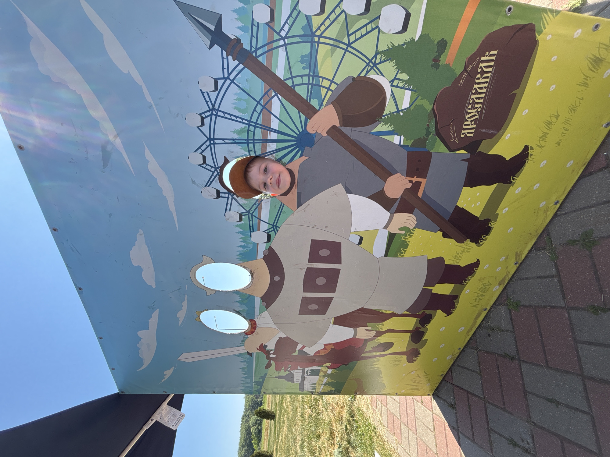 A young boy with brown hair and a brown baseball cap is posing for a photo in a cutout board at an outdoor amusement park or fairground. The board depicts two cartoonish figures, one on horseback and another standing warrior, with cutouts for faces. The boy's face is visible through the cutout in the standing warrior's figure. He is holding a long spear. Behind the figures, the background features a Ferris wheel and stylized trees, under a bright blue sky with white clouds. The foreground shows patterned paving stones, with a shadow cast by the cutout board. The scene appears to be located in or near the city of Zavolzhye, Russia, given the presence of a stone with Cyrillic inscription that translates to "Zavolzhye".