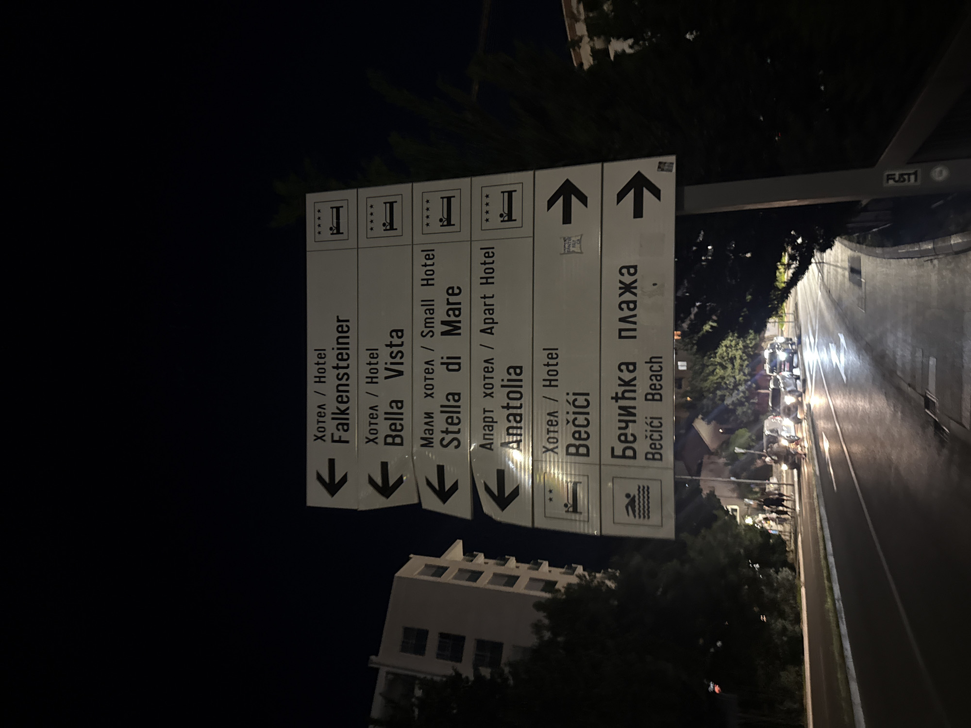 In Rafailovići, Montenegro, at night, a directional signpost stands by a road. The signpost, made of white panels with black text and symbols, displays directions to various hotels and a beach. The hotels listed are Falkensteiner, Bella Vista, Stella di Mare, and Anatolia, each indicated with a left arrow and a hotel icon with four stars. Below these, a sign for Bečići Beach shows a swimming icon and a right arrow, with the text in both Cyrillic and Latin scripts. In the background, a modern, multi-story building is visible, along with trees lining the road. Several cars are driving on the wet road, their headlights illuminating the scene, suggesting recent rain. A few pedestrians are walking on the sidewalk. The sky is dark, indicating nighttime.