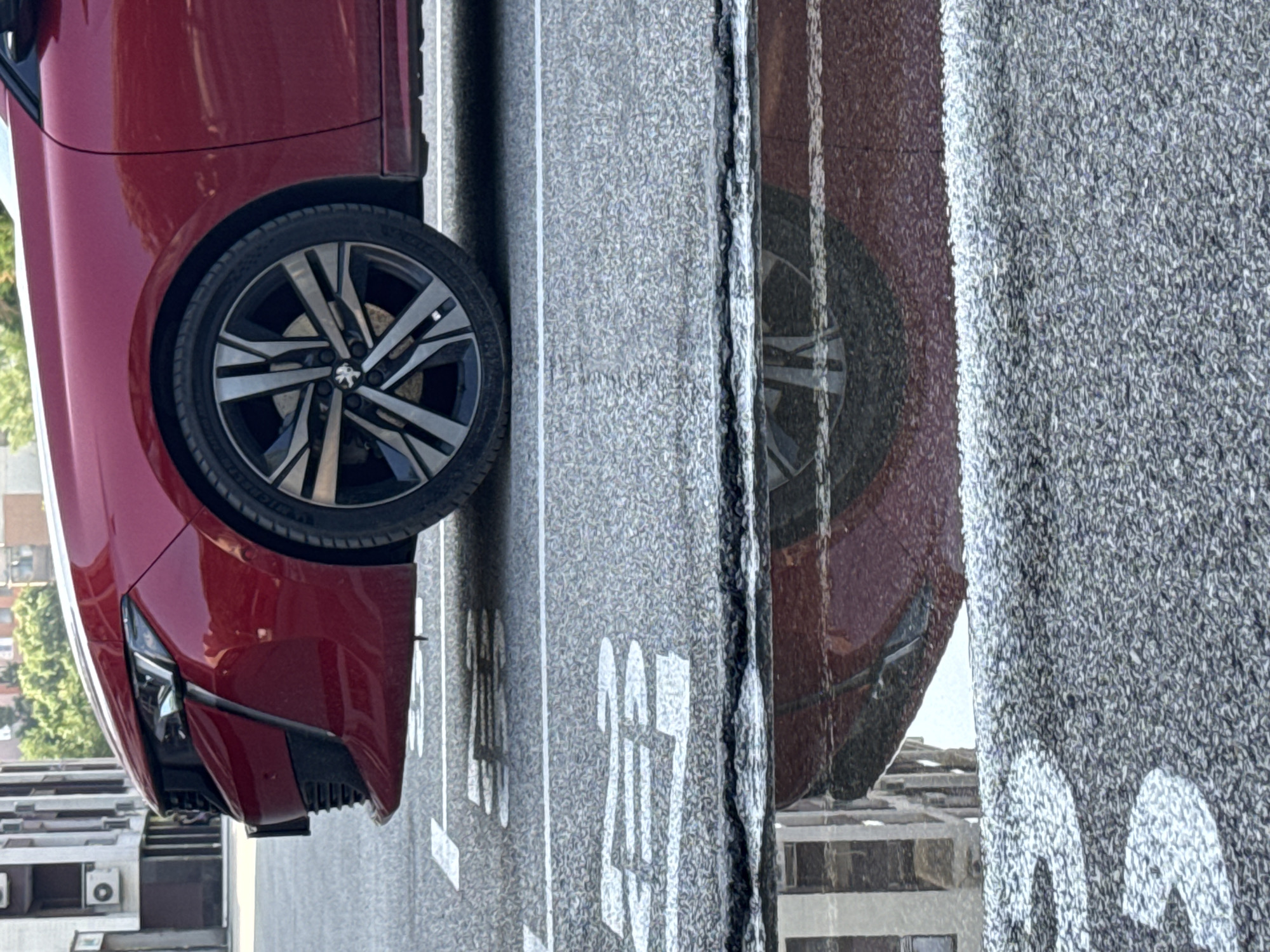 A close-up, low-angle shot captures the front wheel and lower portion of a red Peugeot sedan parked on an asphalt surface. The car's wheel, featuring a multi-spoke alloy rim, is prominent. White painted road markings, resembling parking spot indicators, are visible on the asphalt. A puddle of water in the foreground reflects the car and the surrounding urban environment, including buildings with visible windows and architectural details. The scene appears to be a parking lot or street within a city. The image does not contain any people, and no specific details suggest the location is Plovdiv, Bulgaria.