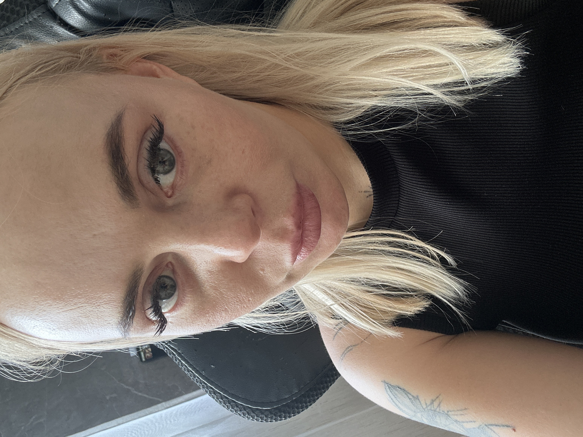 An adult female subject with fair complexion and straight, long blonde hair is depicted in a close-up, head-and-shoulders portrait. She has light-colored eyes with extended eyelashes, dark, well-defined eyebrows, and her lips are slightly parted with a neutral expression. She wears a black ribbed top, appearing sleeveless or with very short sleeves. A faded, light-colored tattoo, resembling a feathered or organic motif, is visible on her upper left arm. A small, dark, indistinct tattoo mark is also present on her lower neck/upper chest area. She is seated in a dark, textured high-backed chair, facing the camera directly. The background is an out-of-focus indoor setting, featuring a light-colored vertical element (possibly a curtain or blind) on the left and darker, indistinct furniture or walls further back. The location is Unknown, Unknown.