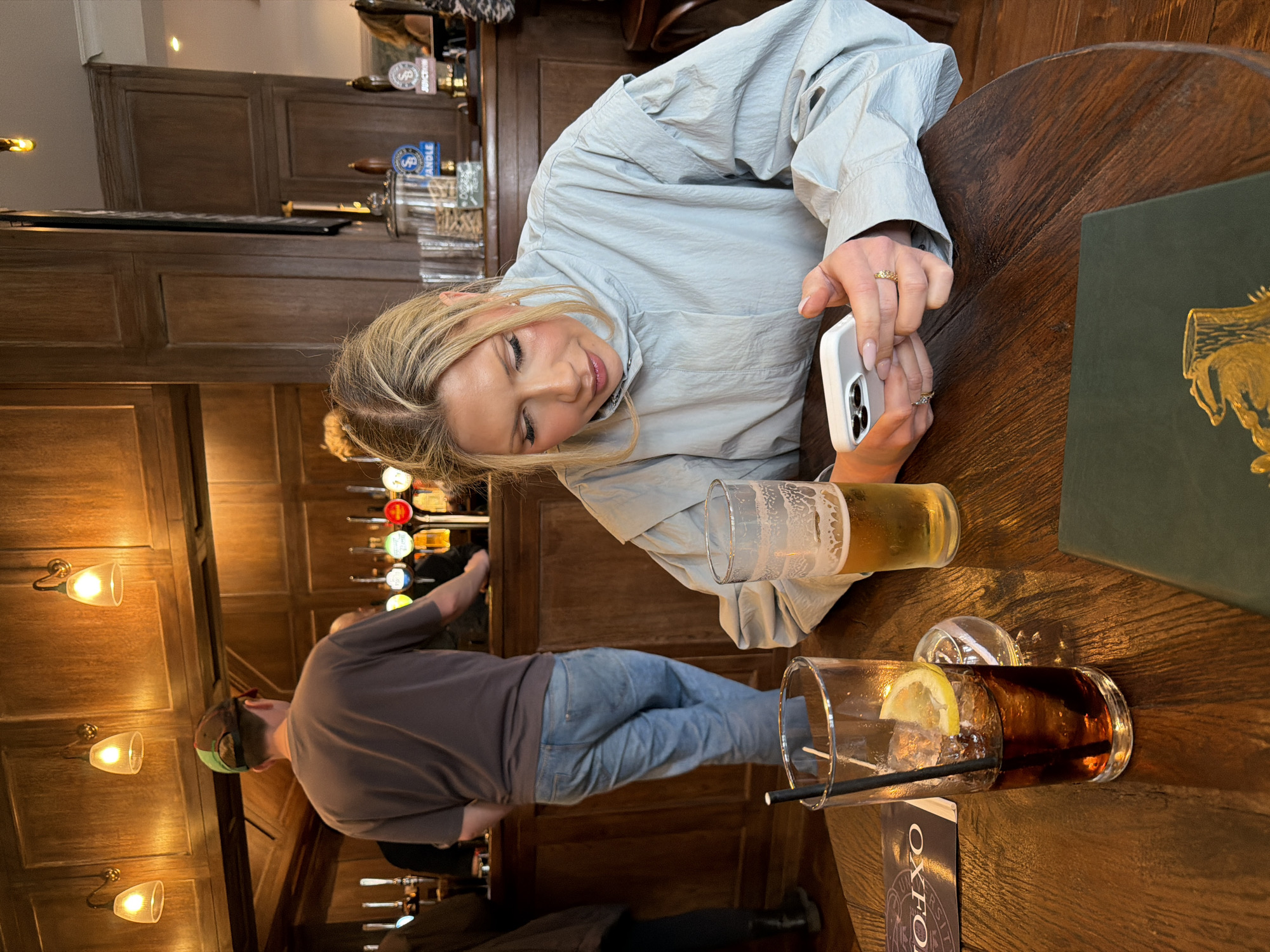 The image depicts a woman at a wooden table in what appears to be a pub. She is holding a smartphone and looking at it. On the table are two drinks: a half-full glass of beer and a glass of dark liquid with ice and a lemon slice, along with a straw. A dark green menu rests on the table. In the background, a bartender is visible behind a bar with multiple beer taps, suggesting a traditional pub setting. The name "OXFO" is visible on an object on the table, possibly indicating the location is in or related to Oxford. The setting seems to be in a traditional pub environment in London, United Kingdom.