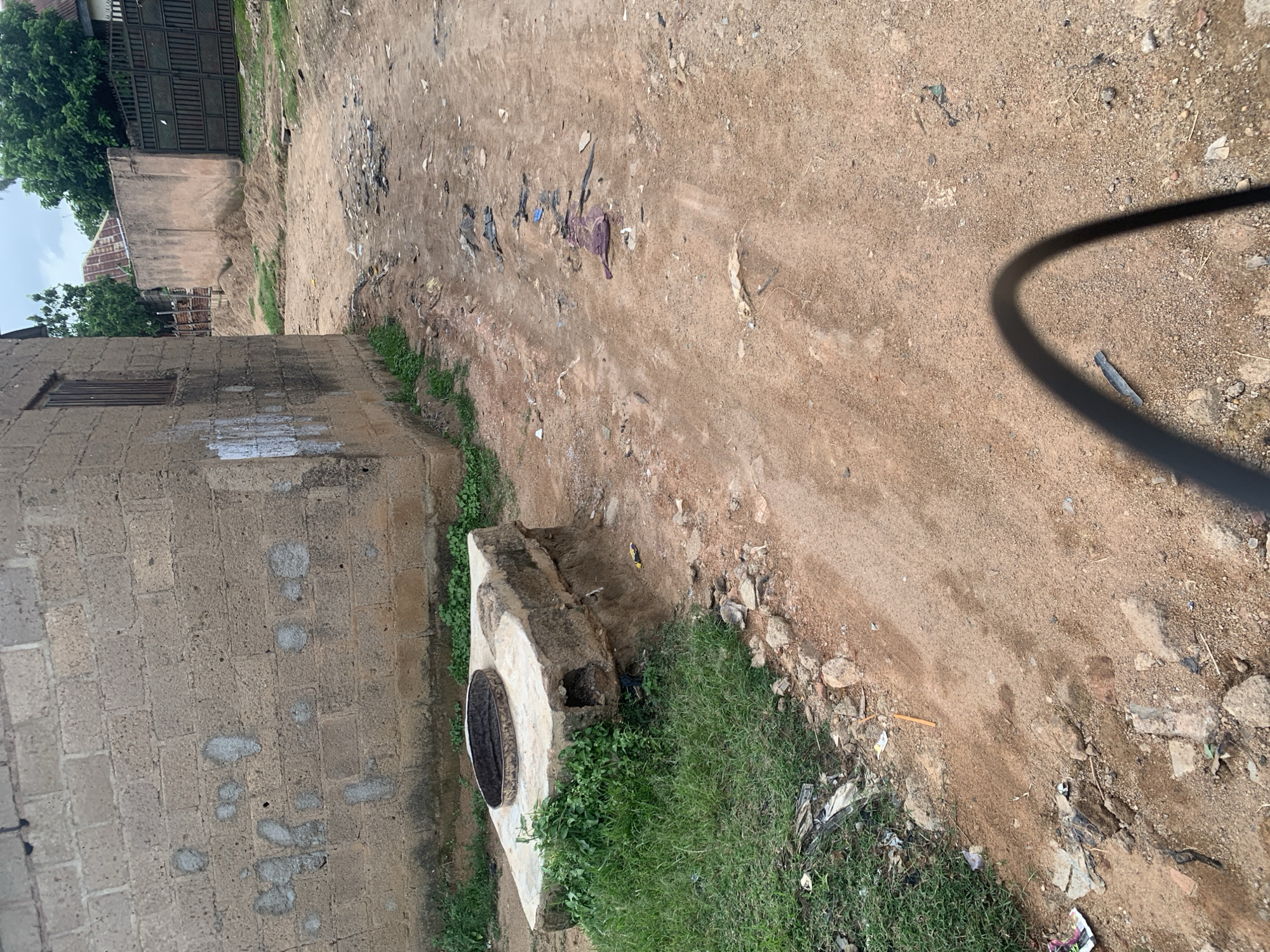 The image depicts a dirt road in Jos, Nigeria, lined with a cinder block building on the left and various debris scattered across the ground. In the foreground, to the left, is a concrete structure with a circular opening, possibly a well or drain, partially overgrown with grass. The building, constructed from unpainted cinder blocks, has a single window with bars and some white markings on its wall. The road itself is unpaved and appears to be a common pathway, with tire tracks visible in places. Further down the road, there are sand piles, a metal gate, and other buildings in the background, suggesting a residential or semi-industrial area. The overall impression is one of a somewhat unkempt urban environment.