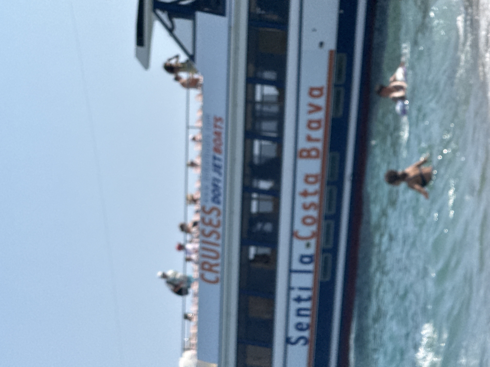 In Blanes, Spain, the image captures a bright, sunny day with a clear blue sky above a body of water. A large boat with "CRUISES" and "DOFI JET BOATS" written on its side is prominent. Below this, the boat's hull displays "Senti la-Costa Brava" in large, orange lettering. Several people are visible on the upper deck of the boat, appearing to be passengers enjoying the cruise. In the foreground, two young individuals are in the water, one on their stomach with a purple inflatable ring, and the other facing away from the camera. The water is choppy with small waves and glints of sunlight.