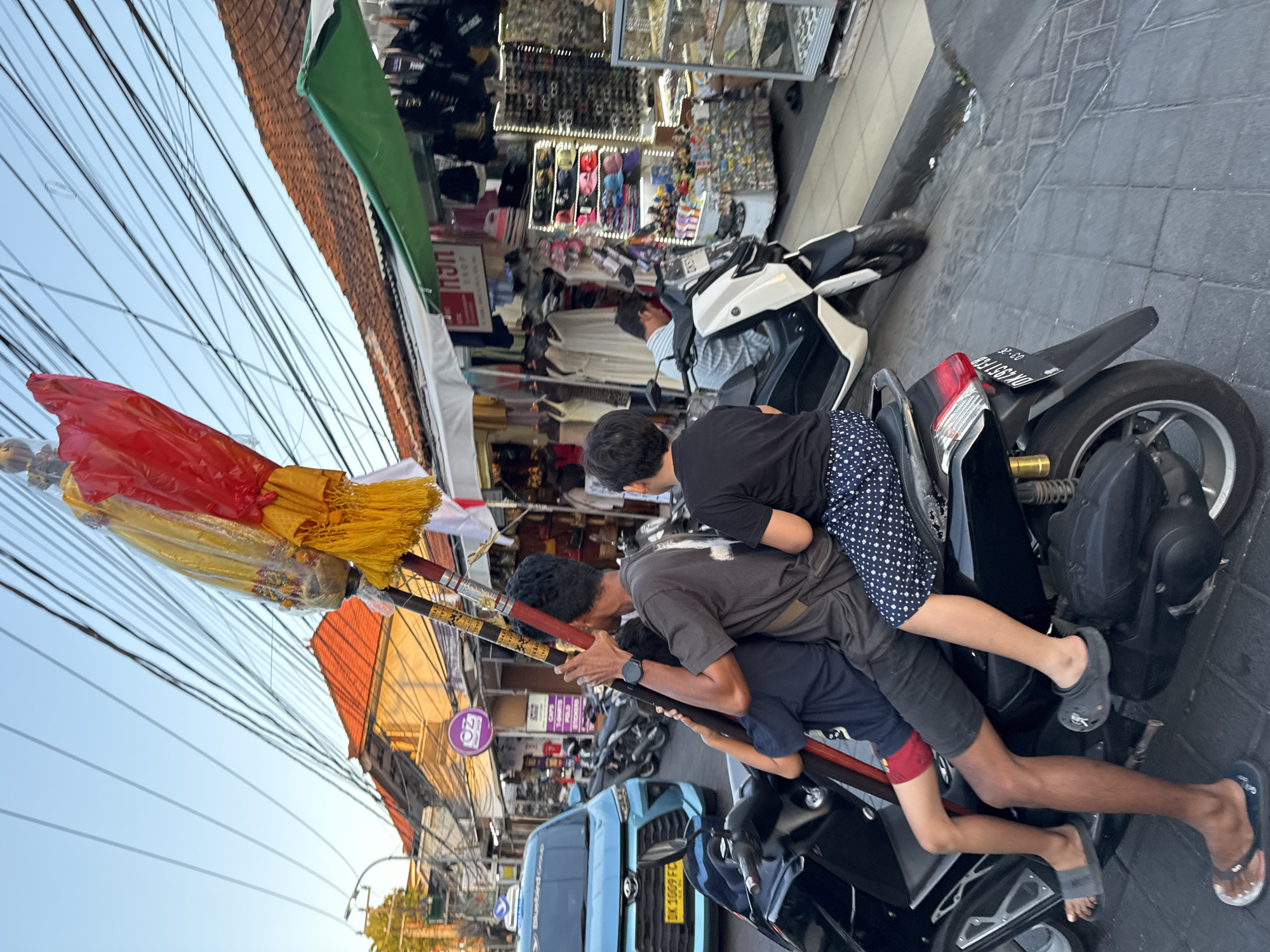 In the city of Kuta, Indonesia, at dusk, three individuals, two children and one adult, are riding a black scooter. The adult is in the middle, with one child in front and another behind. They are carrying a tall, ornate pole topped with red, yellow, and white fabric, possibly a ceremonial banner. The scooter is moving down a street lined with shops selling various goods, including clothing, sunglasses, and souvenirs. The sky is clear blue with a slight orange tint, indicating the setting sun. Electrical wires crisscross overhead, a common sight in Kuta. A blue taxi is visible in the background, along with other parked scooters. The pavement is made of interlocking gray bricks.
