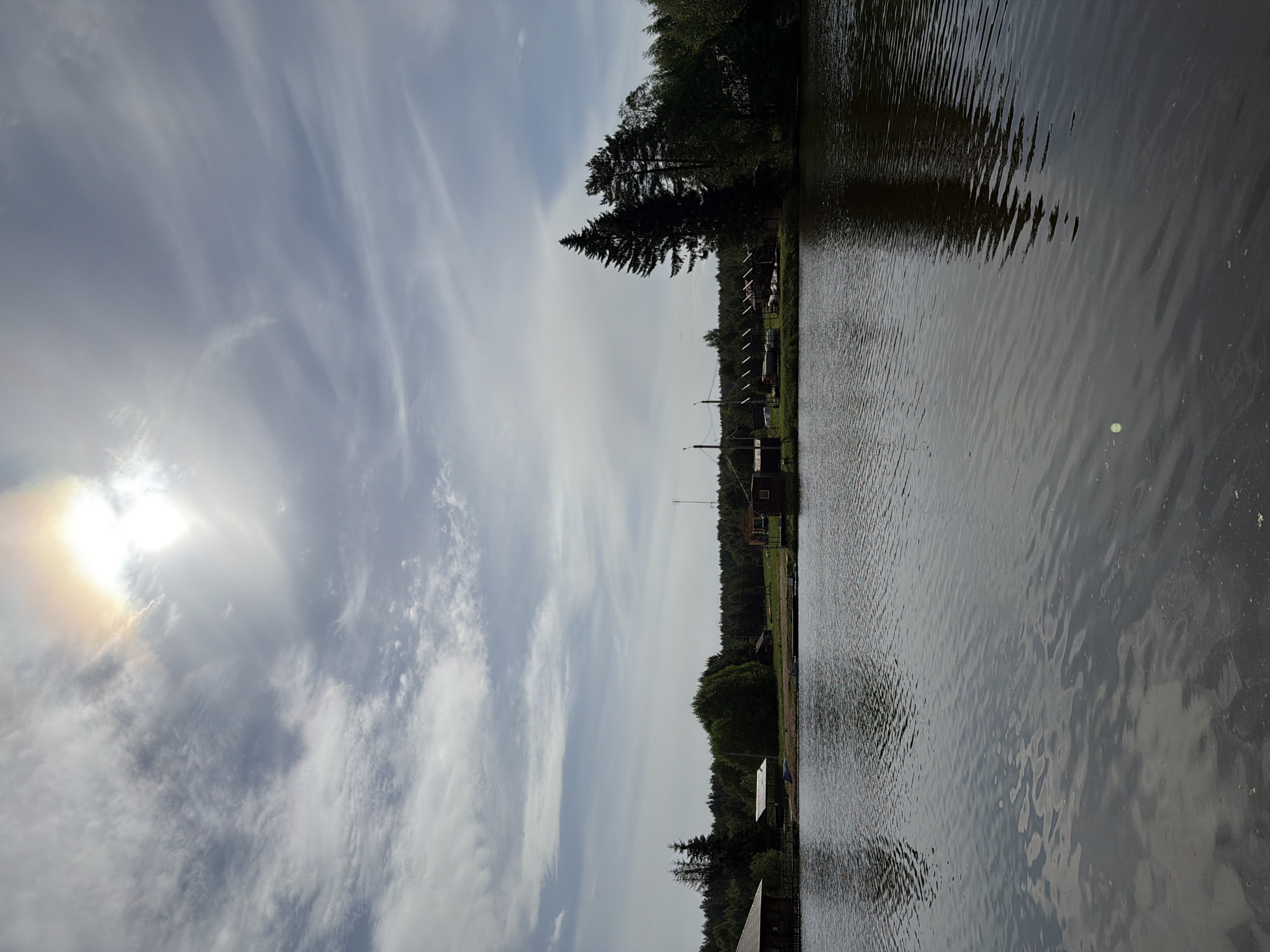 This image shows a body of water, possibly a lake or river, adjacent to a wooded shoreline. The water surface is rippled, reflecting the sky. The sky is mostly overcast with some breaks showing the sun with a halo effect. Along the shoreline are trees and what appears to be a structure. The location is Prikamskii, Russia, based on the provided context.