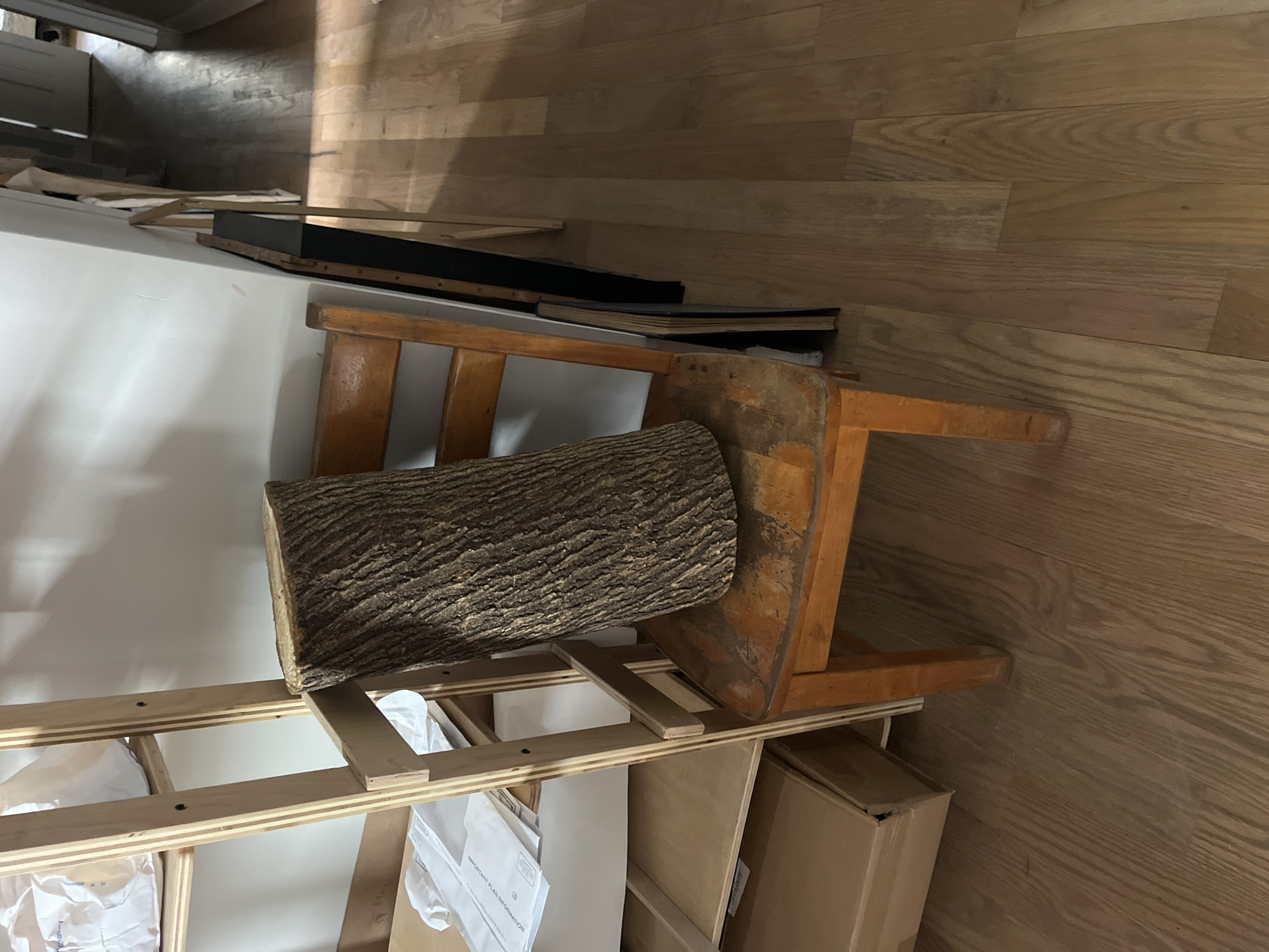 The image depicts a jumbled collection of items in what appears to be an indoor storage or work area. The primary focus is a large, rough-barked log resting on a wooden stool. The stool itself is aged and appears to be made of light-colored wood with a warm, orange-toned finish, showing signs of wear. Behind the stool, a large, flat white surface is visible, possibly a piece of material or a wall.
Further back, several stacked items are discernible. These include what looks like a black frame or box, and some books or binders with dark covers. To the left and below, a light-colored wooden shelving unit is visible, with white materials and documents placed within its compartments. Some of these documents have visible text, including the words "IMPORTANT PLAN INFORMATION." Cardboard boxes are also present in the lower right portion of the image.
The scene is lit from overhead, casting shadows that suggest an indoor environment. The flooring is made of light-colored wooden planks, laid horizontally. The overall impression is one of a cluttered, potentially unfinished or organized-for-storage space. There are no people visible in the image. The time of day and weather are not determinable from the image. The location is identified as New York, United States of America.