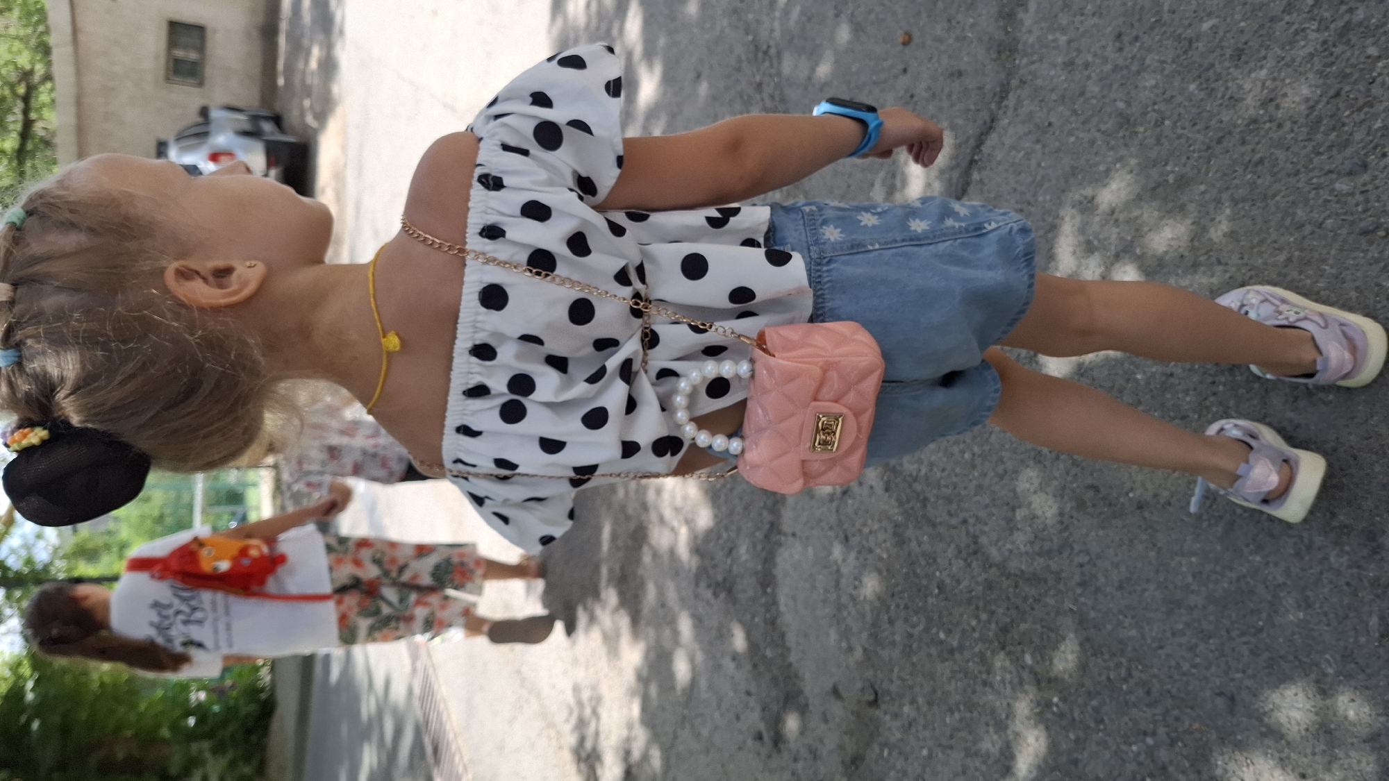 A child walks away from the camera on a paved surface. The child has blonde hair pulled up in a bun with colorful hair ties and wears a white off-the-shoulder top with black polka dots, denim shorts, and light purple sneakers. A small, pink, quilted purse with a pearl handle is slung across the child's shoulder by a gold chain. The child also wears a blue digital watch on their left wrist and a thin yellow necklace. In the background, another child, seen from behind, wears a white t-shirt and patterned pants and carries a red backpack. A silver SUV is parked on the right side of the frame. The environment appears to be an outdoor public space with trees and a building visible. The image was taken in Shymkent, Kazakhstan.