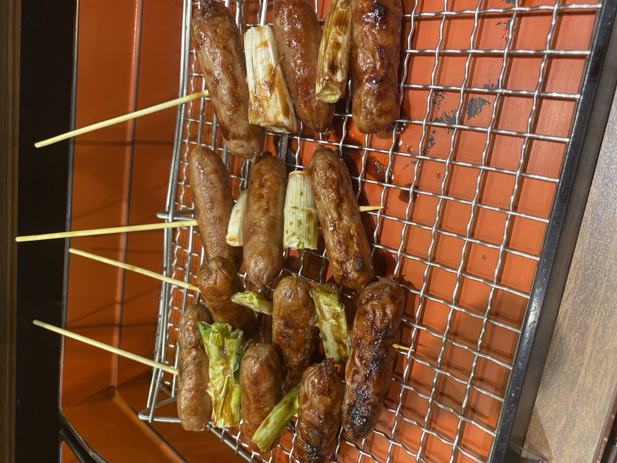 In the city of East Jakarta, Indonesia, on a metal grill, skewers of grilled sausages and scallions are arranged. The sausages are browned and glazed, some with visible grill marks. The scallions are chopped into segments, appearing tender and slightly charred. The grill sits on an orange tray, with a wooden surface visible in the background. The lighting is warm and appears to come from above.